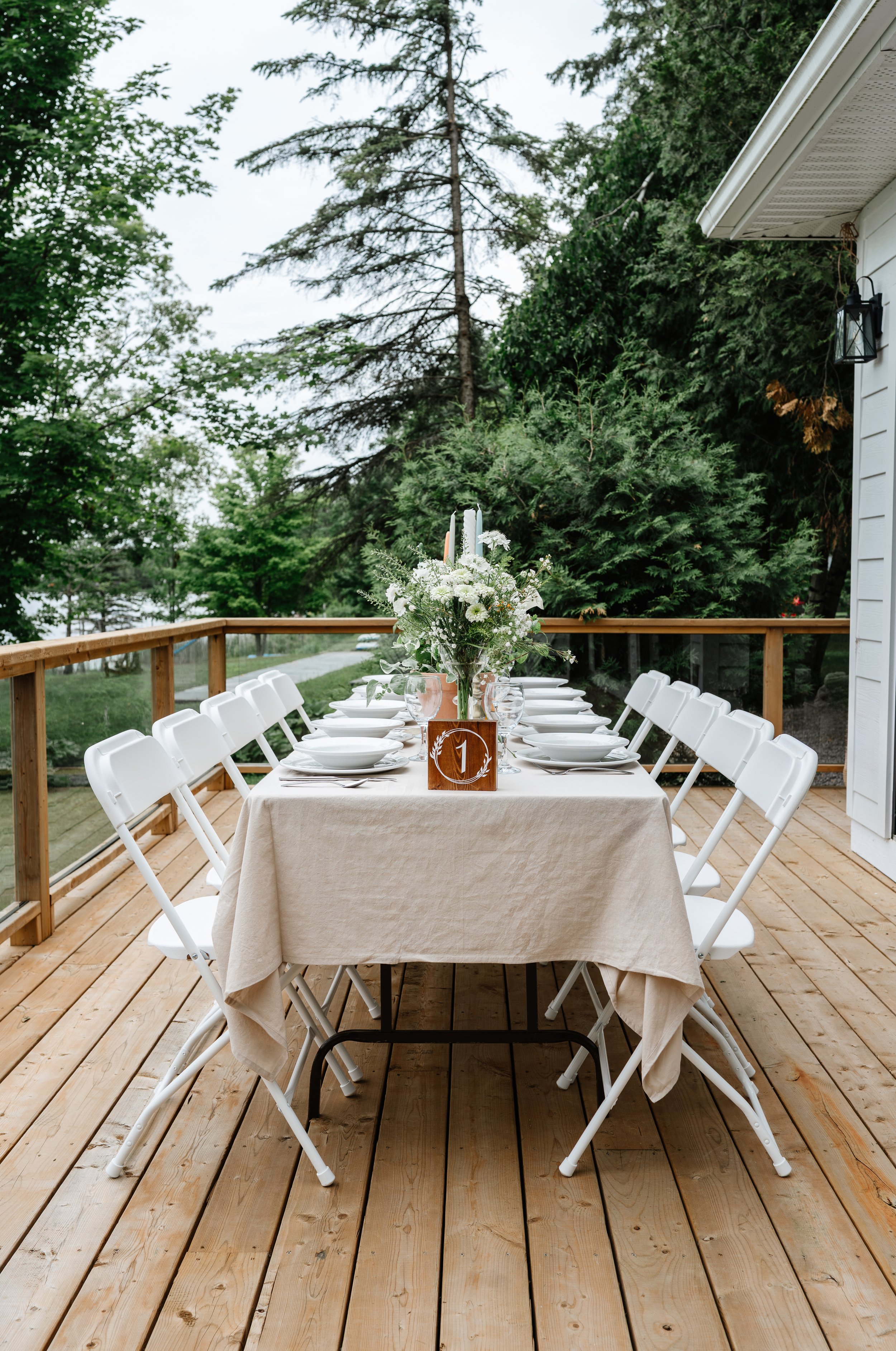 Outdoor dining table set with white chairs, a beige tablecloth, and a floral centerpiece, on a wooden deck with green trees in the background.