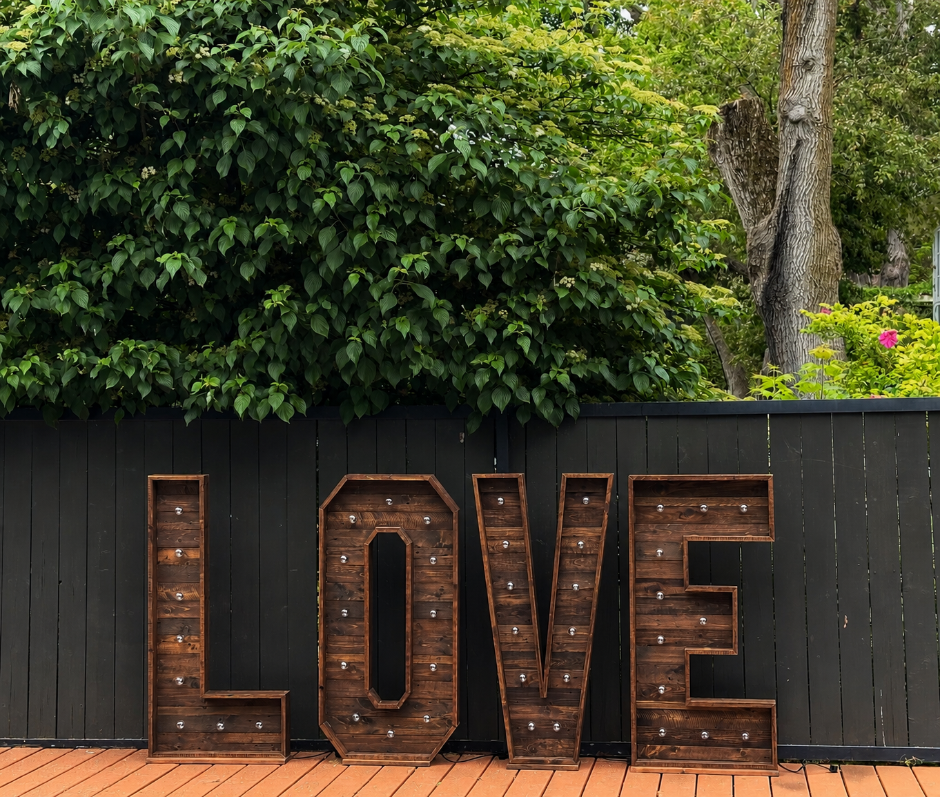 Decorative wooden sign spelling out "LOVE" with lightbulb accents, set against a black fence with green foliage and trees in the background.