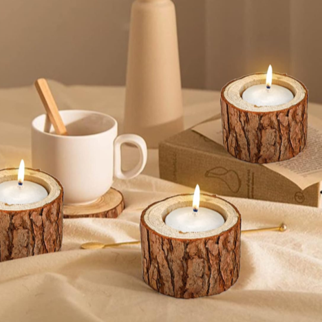 Three lit candles in bark-textured holders and a white mug with a wooden stirring stick on a table with a beige cloth, a book, and a tall beige vase in the background.
