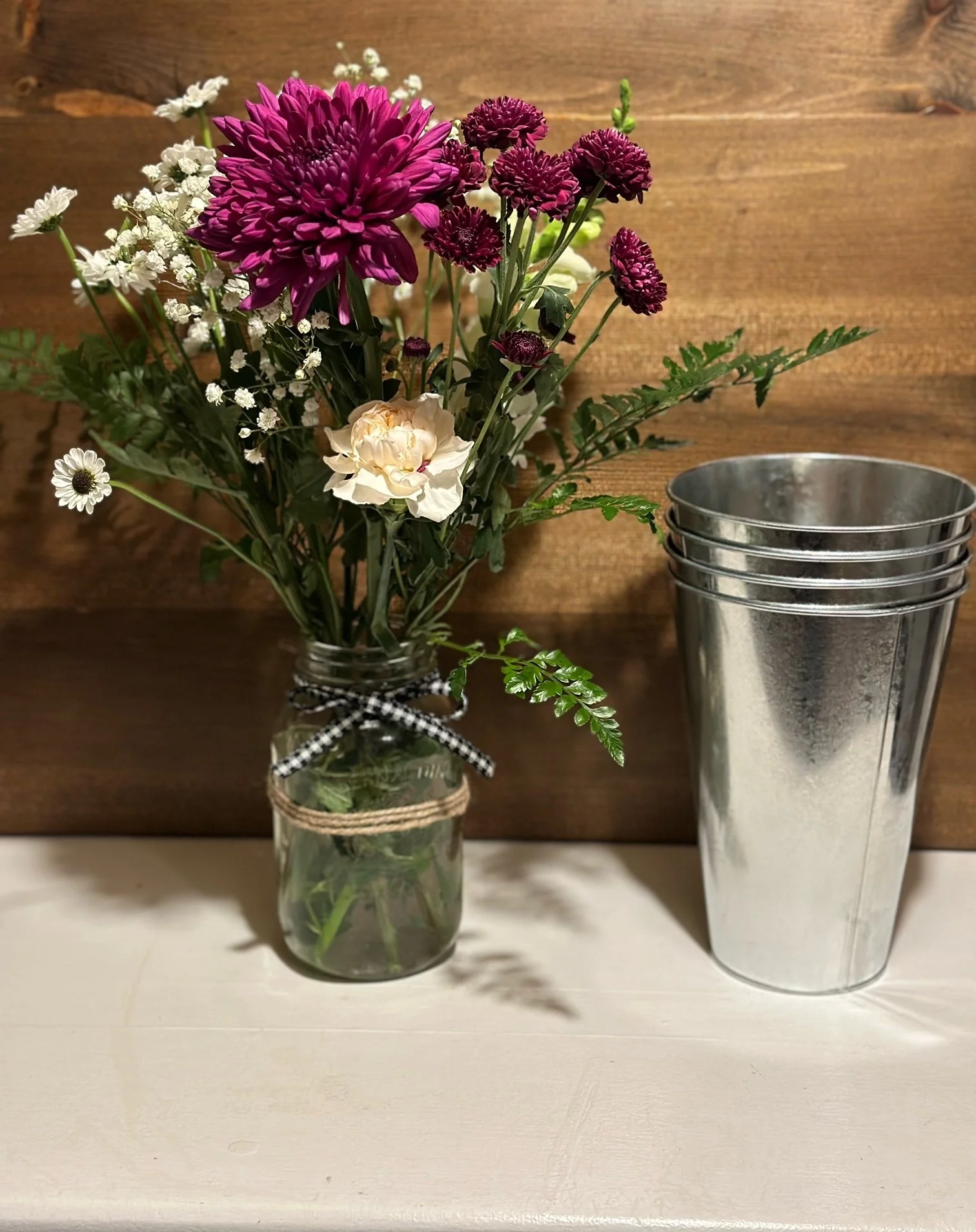 Bouquet of purple, white, and pink flowers in a glass jar on a white surface, with a wooden background and steel tumbler on the right.