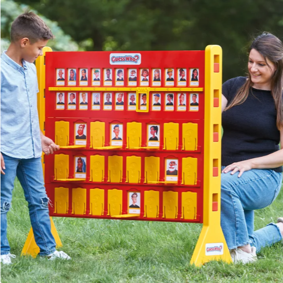 A woman and a boy playing Guess Who outside on grass, with a Guess Who game board displaying various faces.