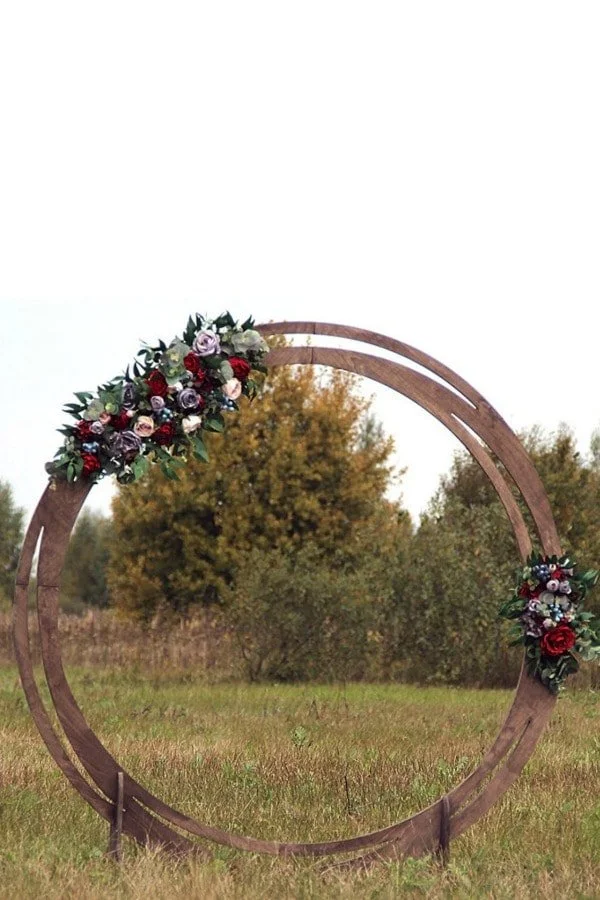 A large circular wooden arch decorated with floral arrangements set outdoors in a grassy field with trees in the background.