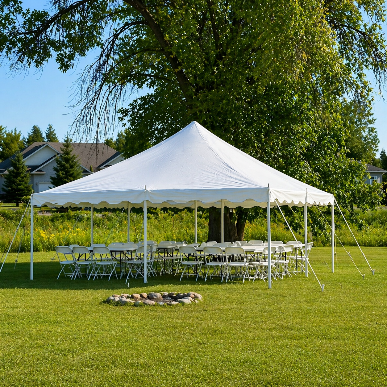 A white event tent set up outdoors on a grassy lawn, with chairs and tables underneath. There is a large tree behind the tent and houses in the background. A small fire pit with rocks is in the foreground.