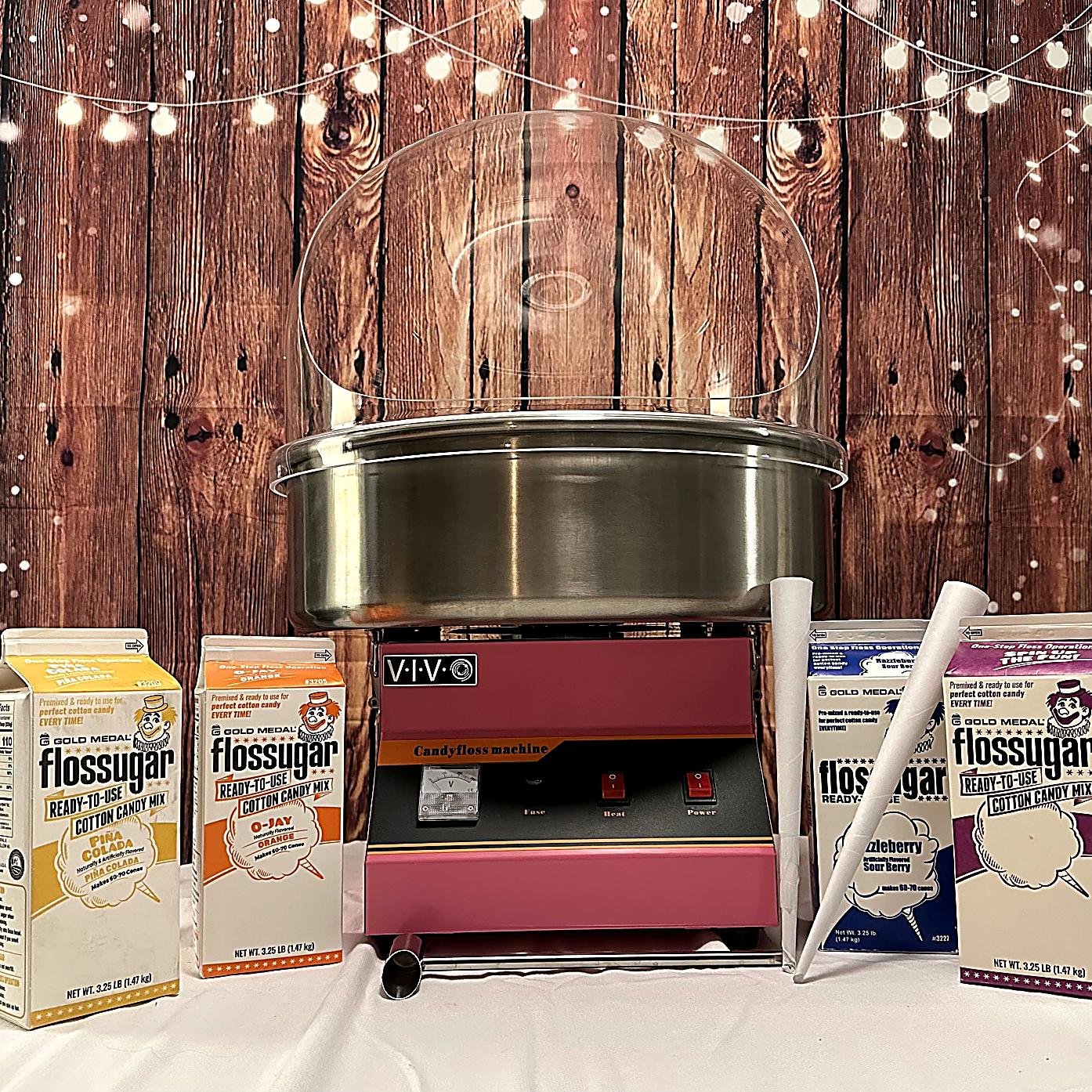 A cotton candy machine with a metal base and a clear plastic dome, surrounded by boxes of Flossugar flavored cotton candy mixes, setting against a wooden backdrop decorated with string lights.