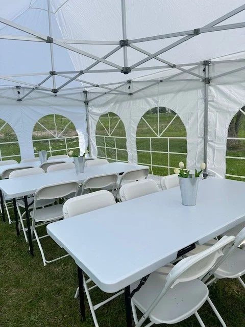 White folding chairs and tables set up inside a white event tent with arched windows, on a grassy outdoor area, each table has a small potted plant.