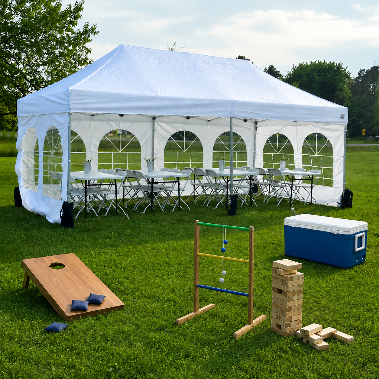 White outdoor event tent with tables and chairs, mini cooler, cornhole game, Jenga, and a wooden bowling game on a grassy field