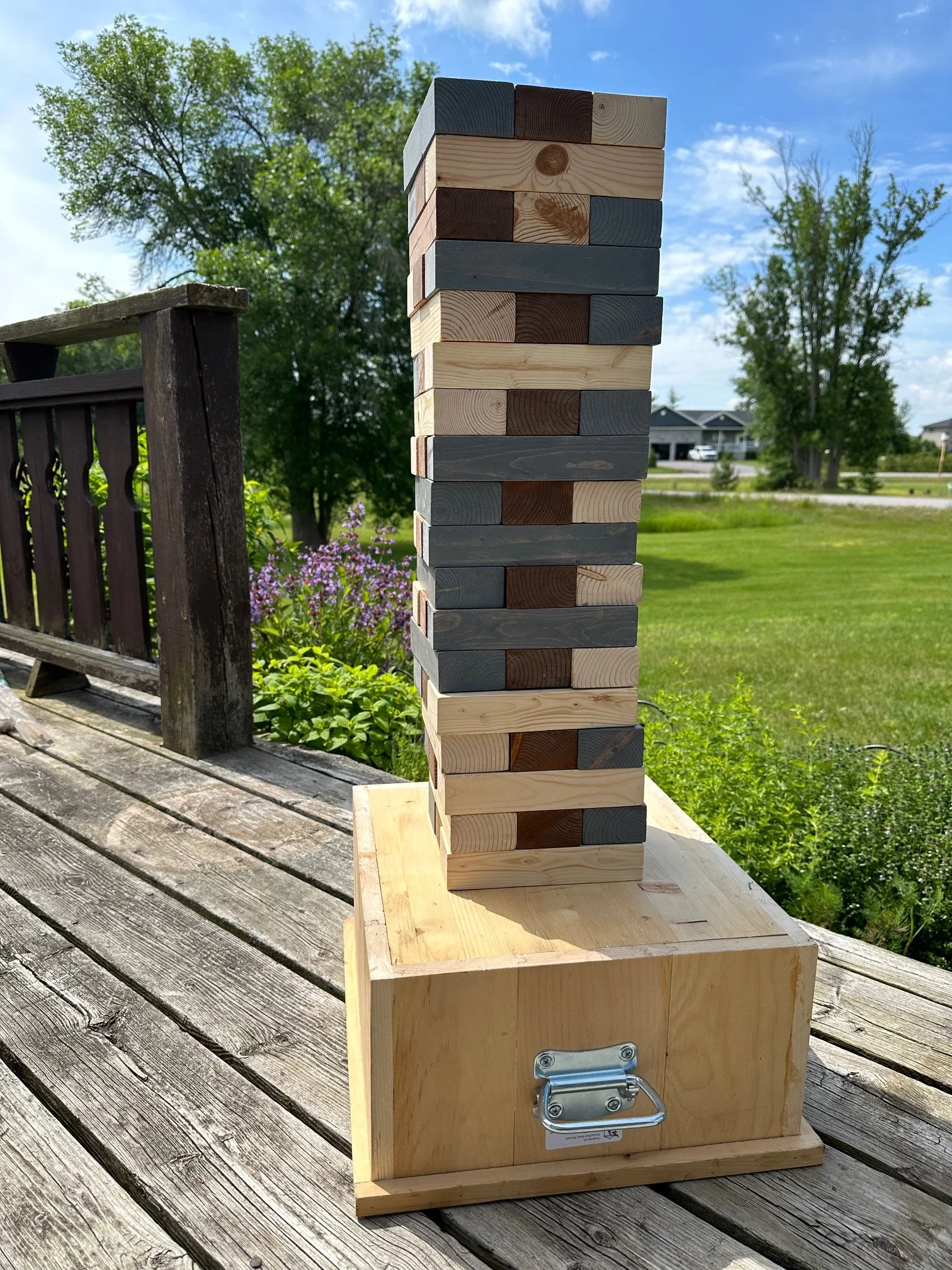 A large wooden block tower game, similar to Jenga, is set up outdoors on a wooden deck, with a wooden box and handle nearby, against a background of trees and houses under a blue sky with some clouds.