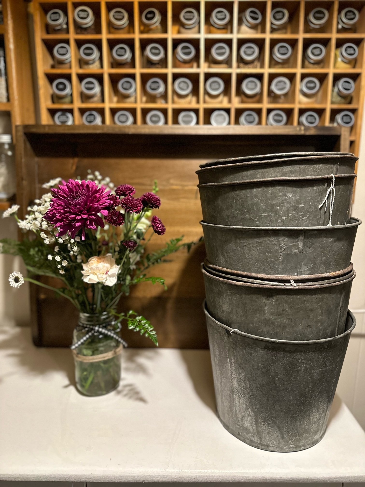A bouquet of flowers in a jar, and four stacked metal buckets on a white surface. In the background, there is a wooden wine rack filled with bottles.