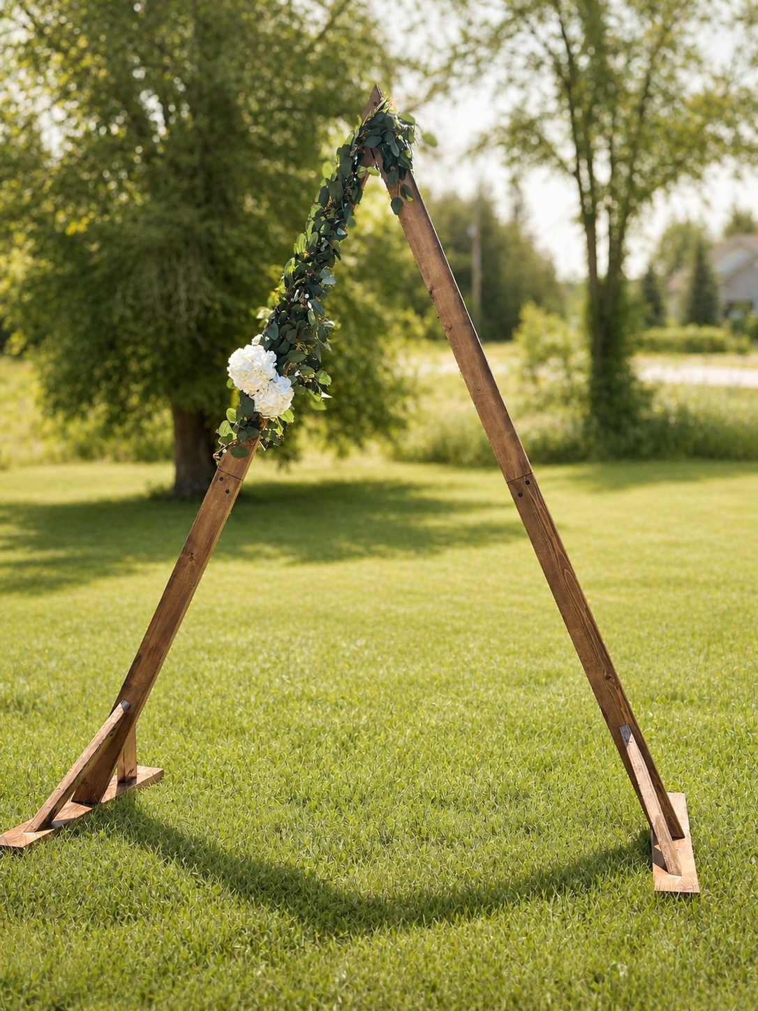 Wooden A-frame stand decorated with green leaves and white hydrangea flowers, set outdoors on grass with trees in the background.