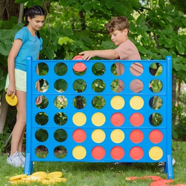 Two children playing Connect Four outdoors surrounded by green foliage.