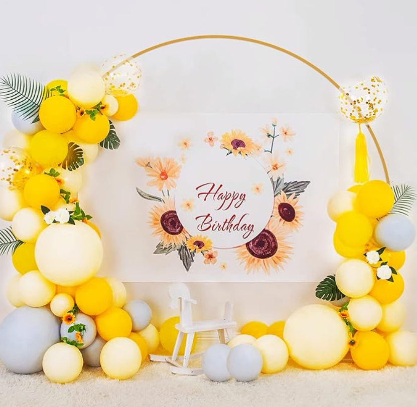 Celebration backdrop with yellow, white, and gray balloons arranged in a semi-circle around a floral sign that reads "Happy Birthday," with a small white rocking horse in front.