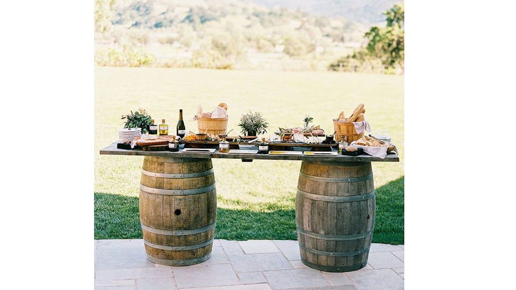 Outdoor buffet setup on a wooden table with wine, flowers, and food, supported by two large wooden barrels in a grassy field.