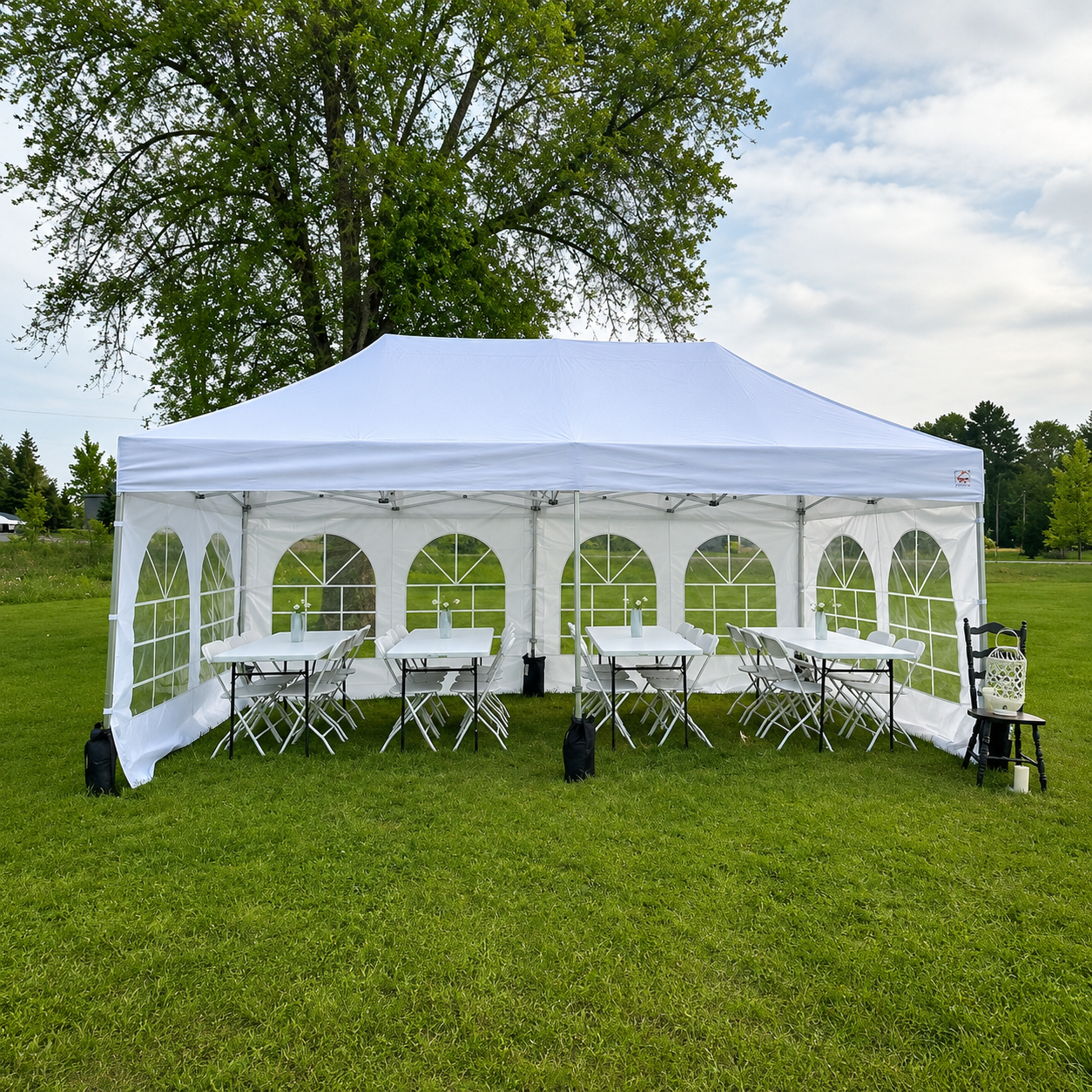 A white event tent set up outdoors on grass with tables and chairs underneath, in a park-like setting with trees and a partly cloudy sky.
