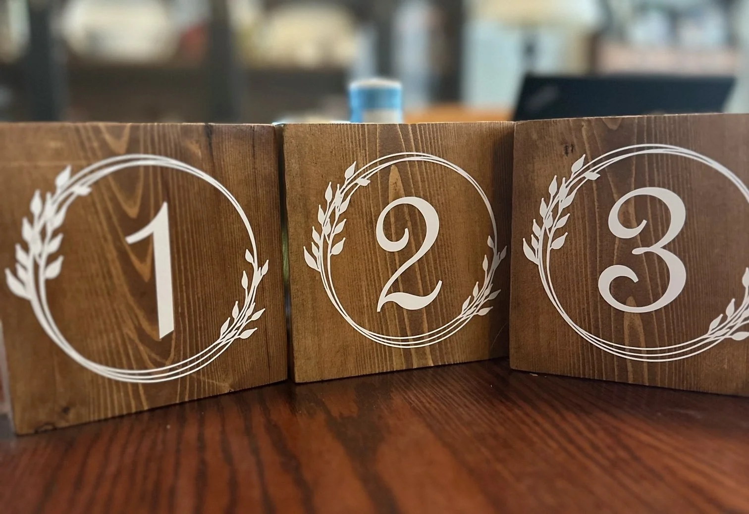 Wooden table numbers 1, 2, and 3 with white circular wreath designs on a wooden surface, used for table seating arrangements at events.