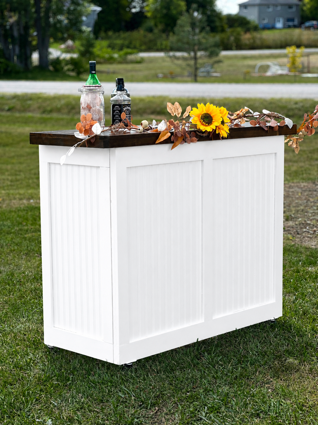 Outdoor bar or counter decorated with a sunflower, gold and white leaves, and a string of small white lights, set on grass with trees and a house in the background.