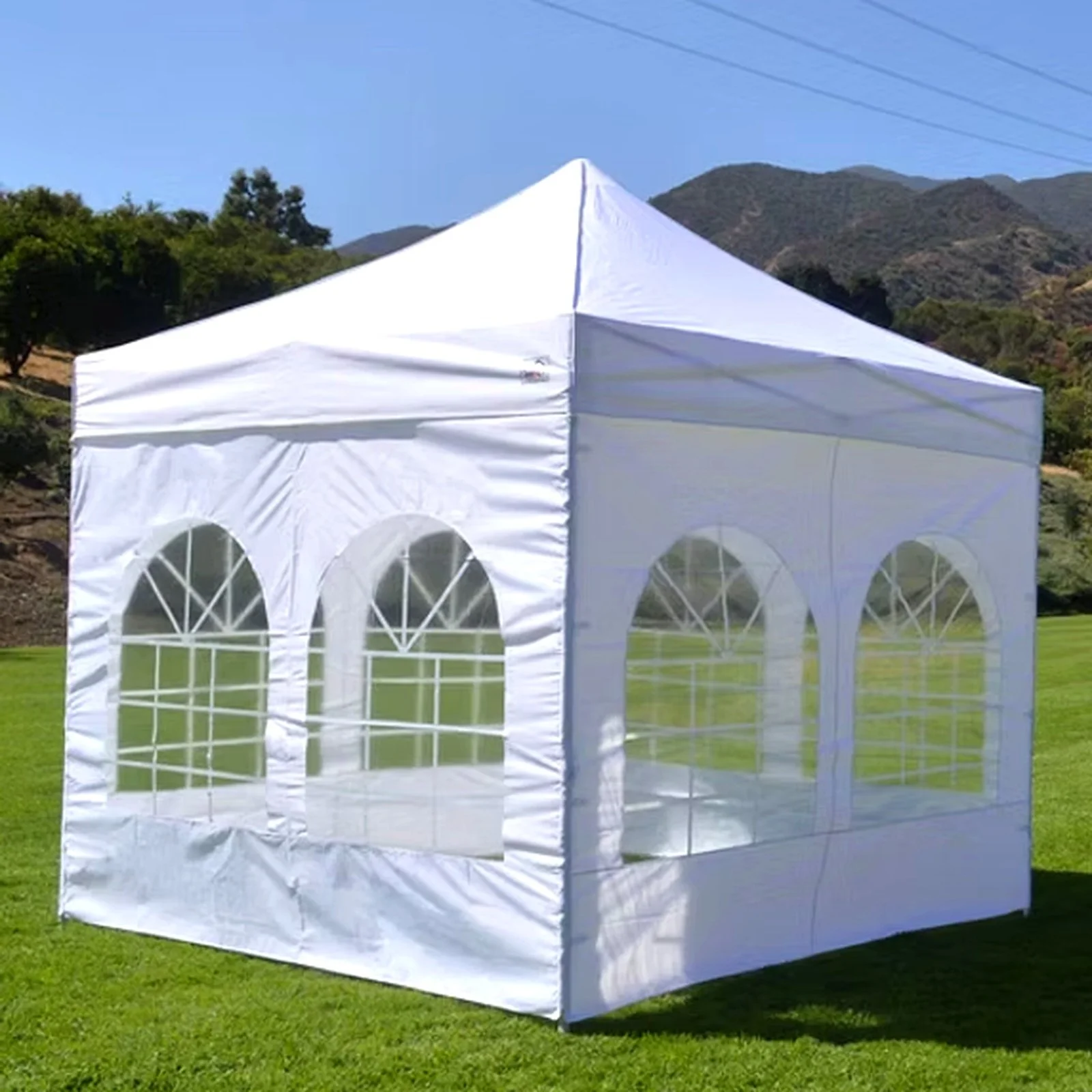 A white outdoor event tent with arched windows set up on a grassy field with hills and trees in the background under a clear blue sky.