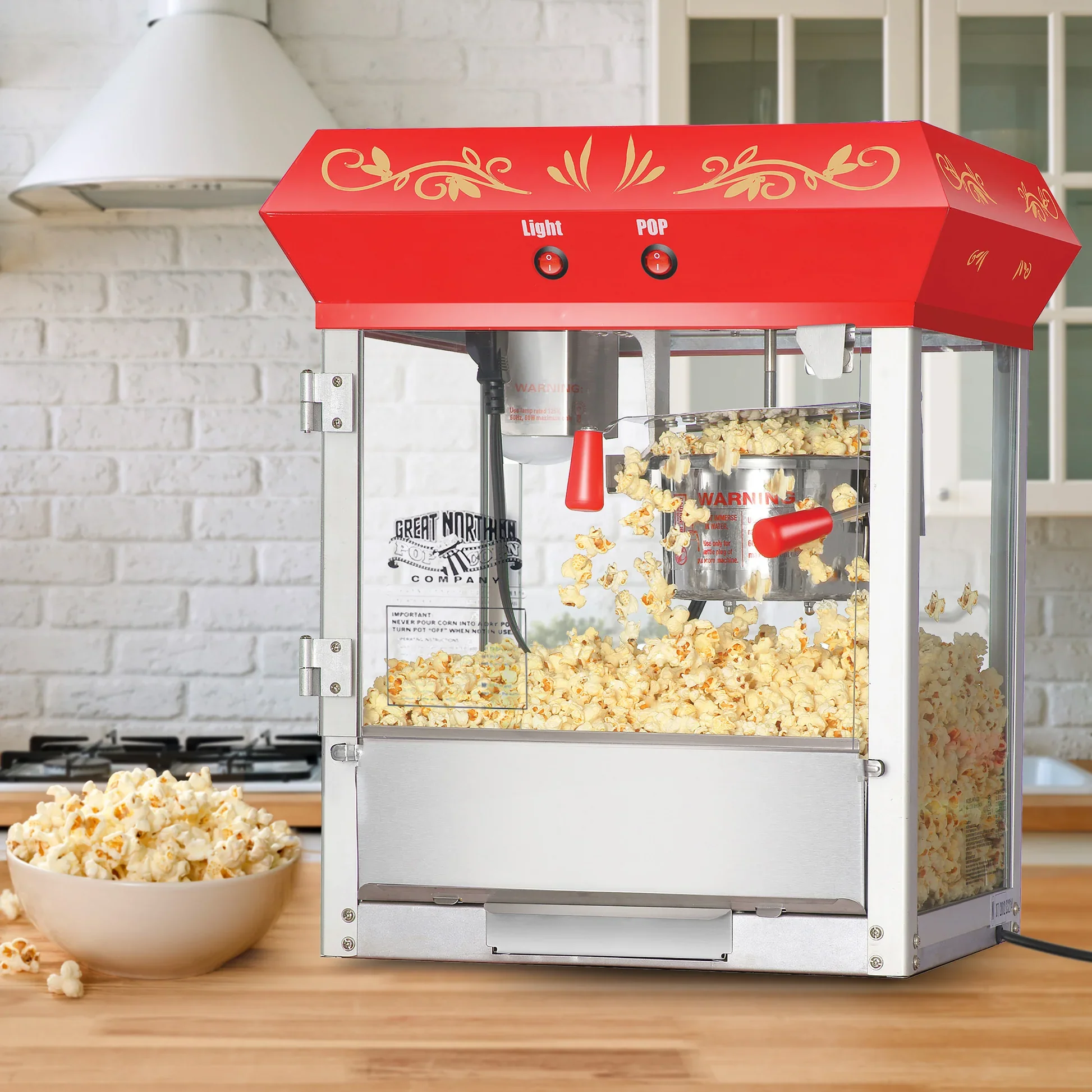 A popcorn maker machine with a red top and gold decorative designs, placed on a wooden table in a kitchen. Popcorn is spilling from the machine into a bowl.