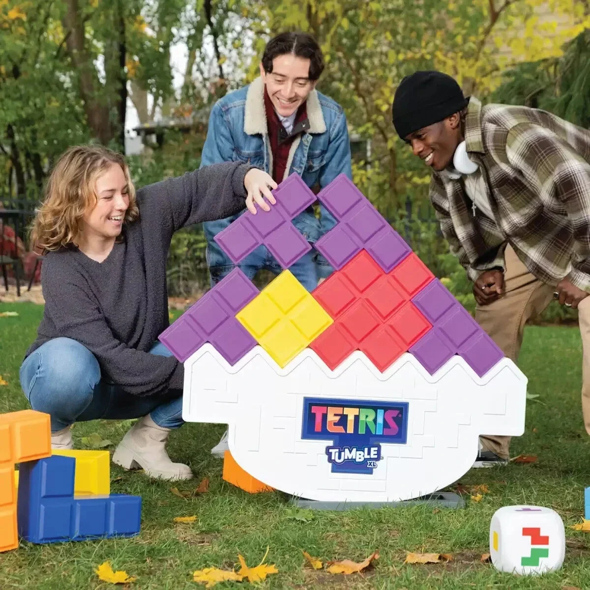 Three people playing Tetris Tumble game outdoors in a park during fall. They are smiling and stacking colorful Tetris-shaped blocks on a large game board shaped like a cupcake.