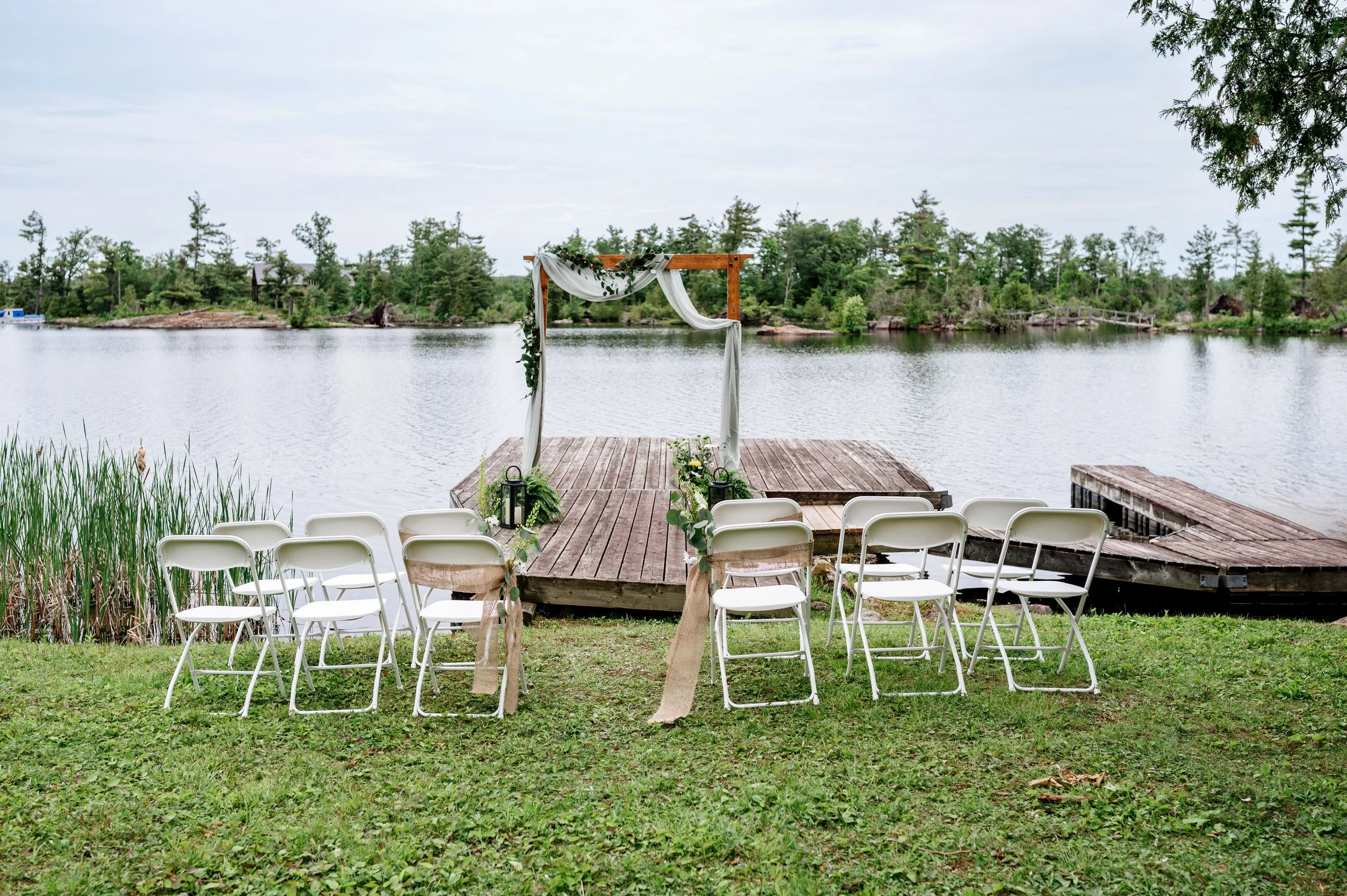 Outdoor wedding setup with white chairs, a wood arch decorated with greenery and white fabric, on a dock by a lake with trees in the background.