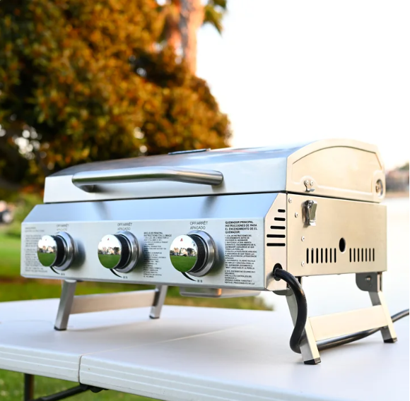 Portable outdoor gas grill with three control knobs on a white table, with trees and a body of water in the background