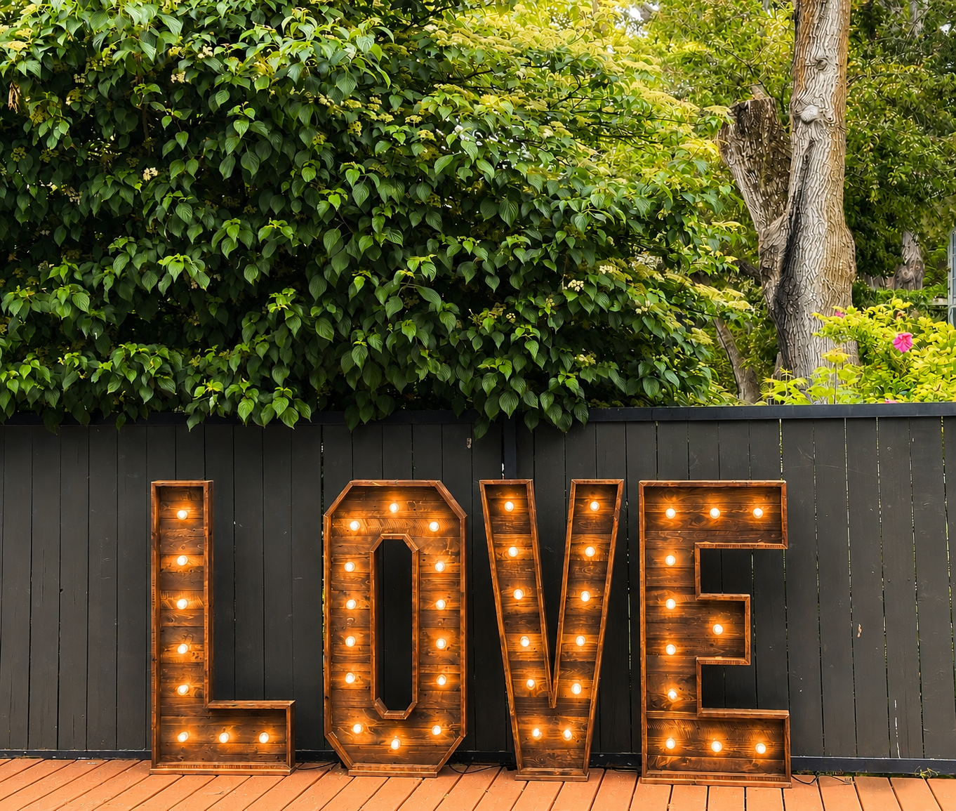 Decorative wooden love sign with illuminated bulbs, placed in front of a dark fence, with lush green foliage and trees in the background.