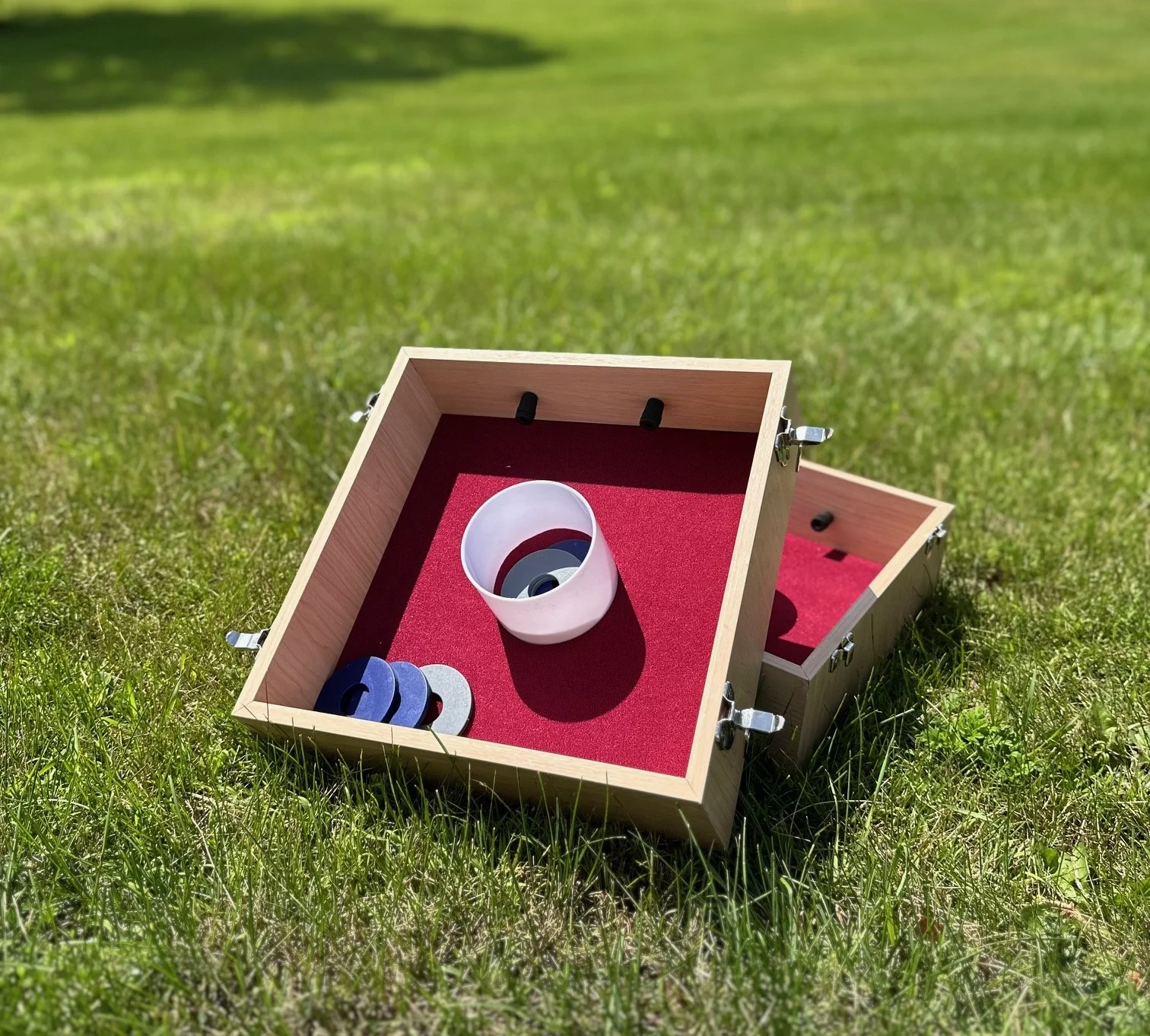 A wooden ring toss game with a red felt interior, placed on a grassy lawn. The game includes colorful rings, with some stacked inside a white bowl and others scattered inside the game box.