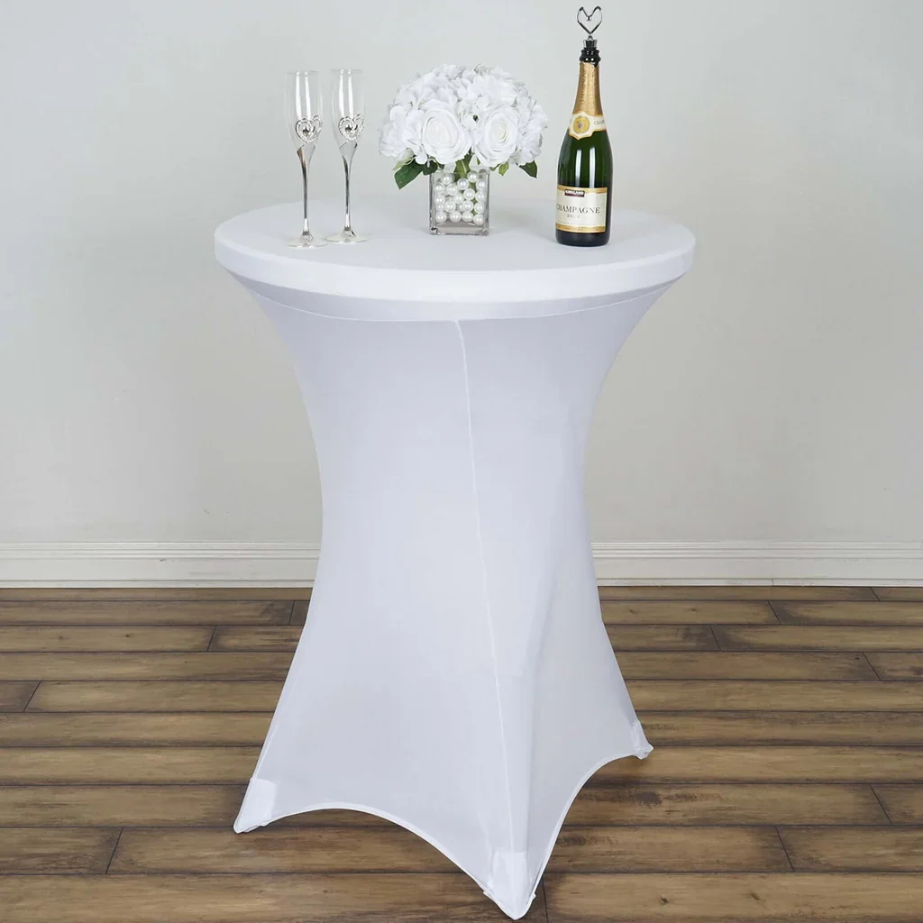 A white cocktail table with a floral centerpiece, champagne bottle, and two champagne glasses against a plain wall and wooden floor.