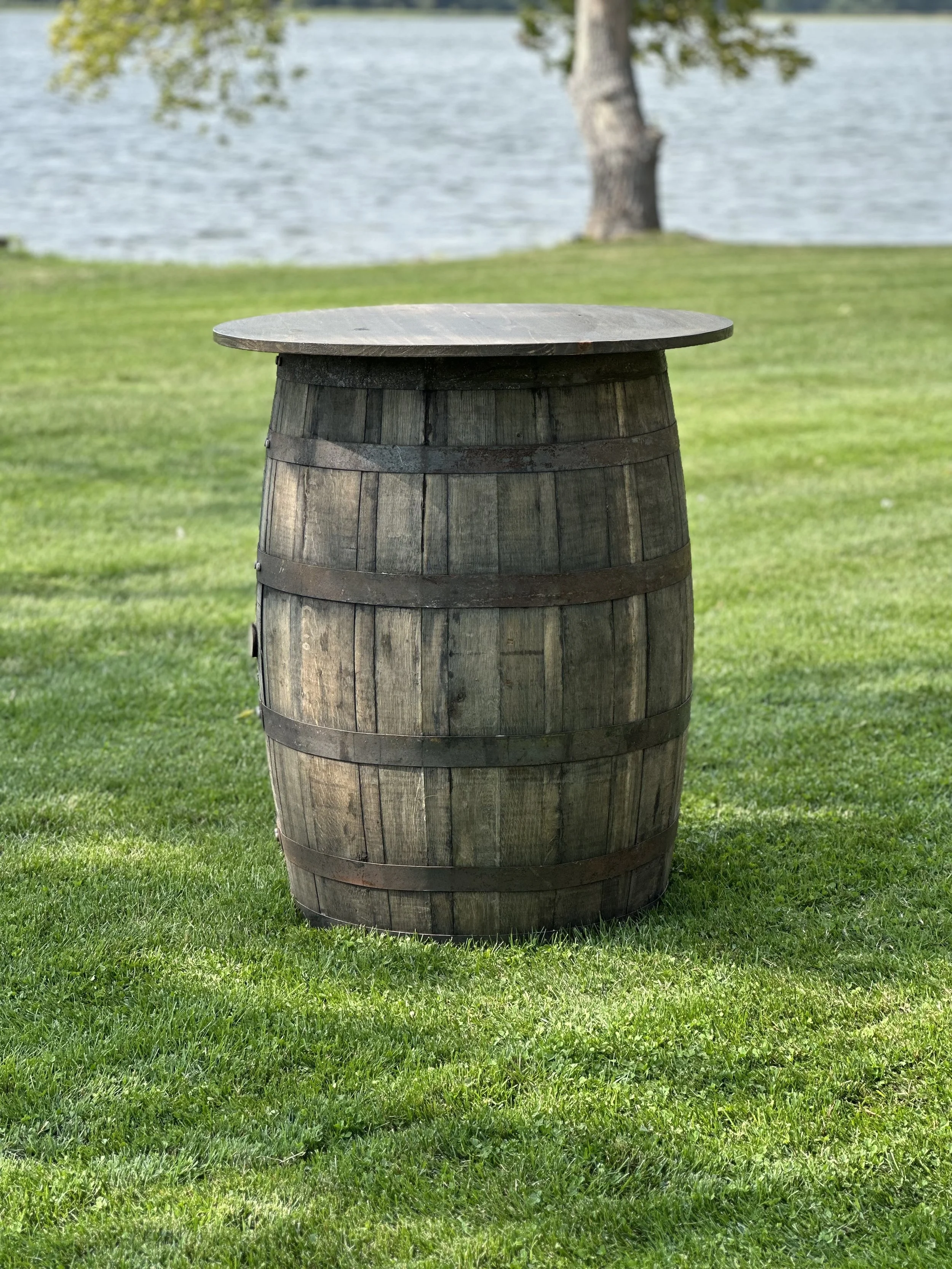 A wooden barrel used as a table outdoors on a grassy area near a body of water with a tree in the background.