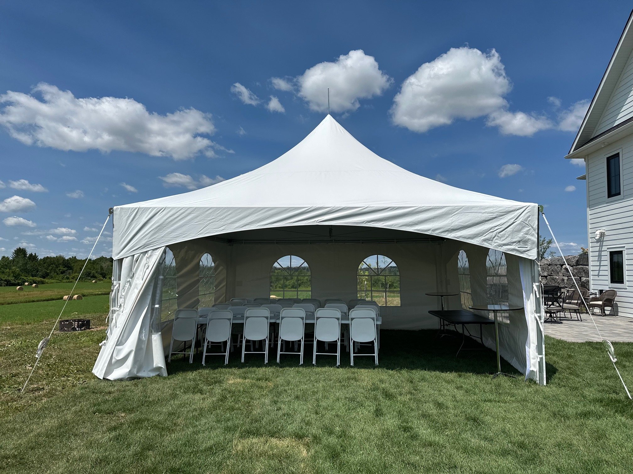 A white event tent set up outdoors on a grassy area under a bright blue sky with scattered clouds. Inside the tent, there are rows of gray chairs arranged in front of a small black table. To the right, part of a white house with black window frames is visible, along with outdoor furniture and a stone wall.