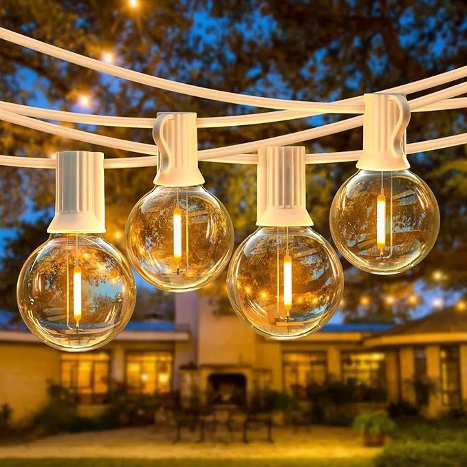 String of four glowing outdoor string lights hanging above a backyard patio, with a house and trees in the background during dusk.