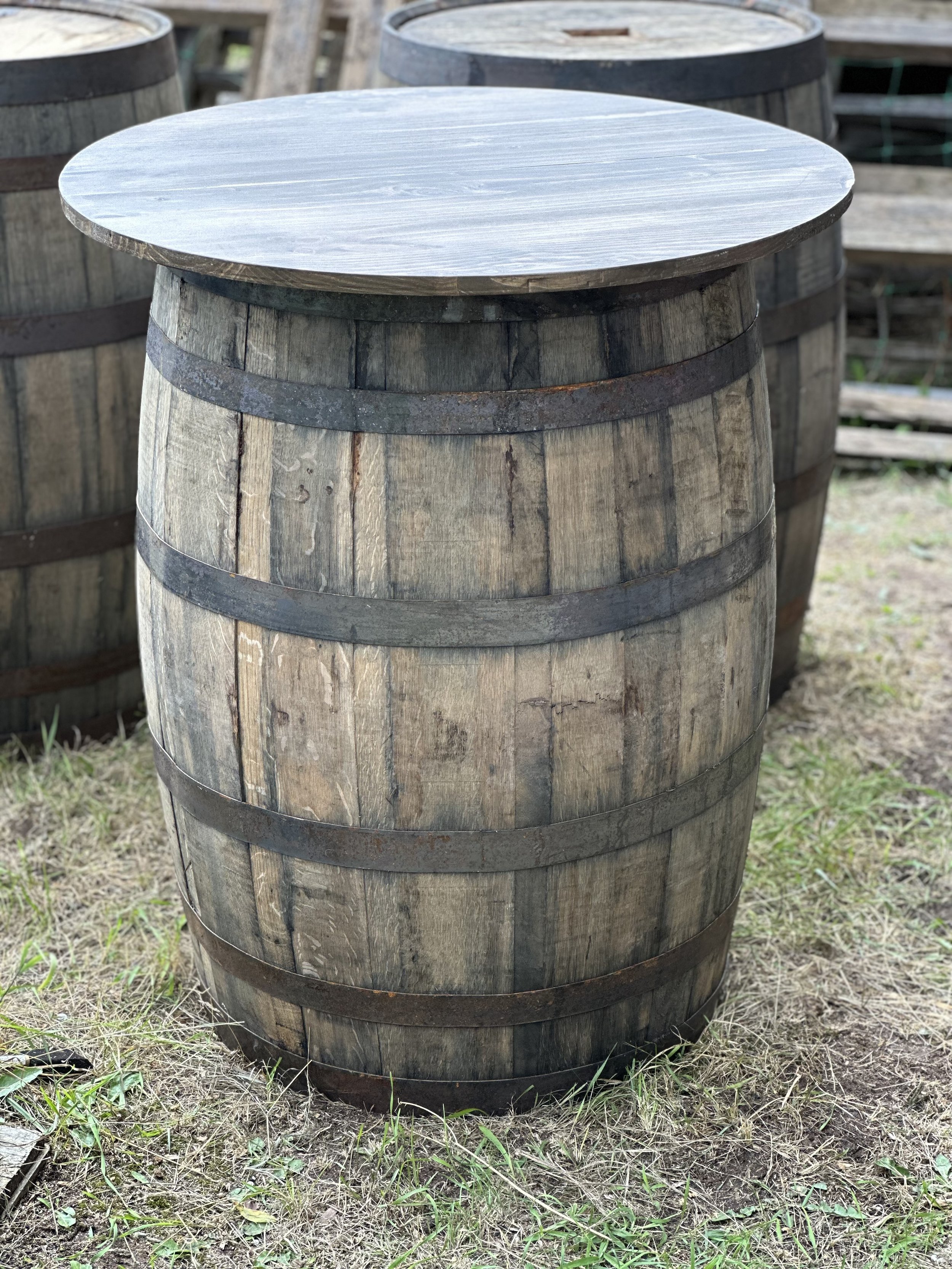 A wooden barrel with metal bands, used as a table, outdoors on grass with other similar barrels in the background.