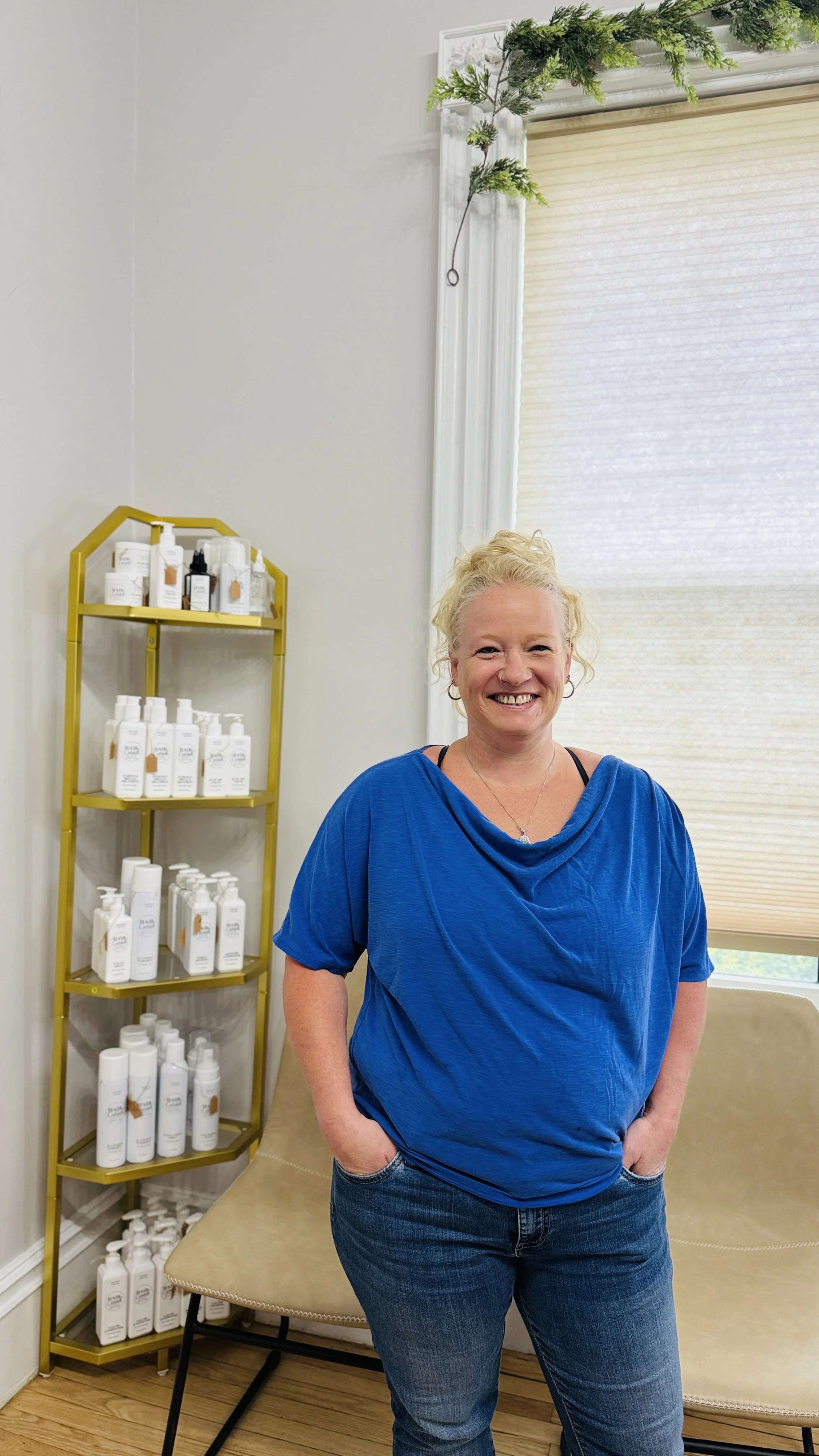 A smiling woman with curly blonde hair wearing a blue top and jeans stands inside a room with a white wall, beige window covering, and a yellow shelf filled with white bottles and containers.