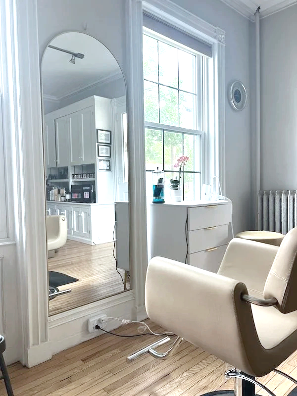 Interior view of a bright room with a large window and white furniture, including a cream-colored chair and a white dresser with a pink flower and decorative items. A tall mirror reflects part of the room, and an electrical outlet with a power strip and cords is visible on the baseboard.