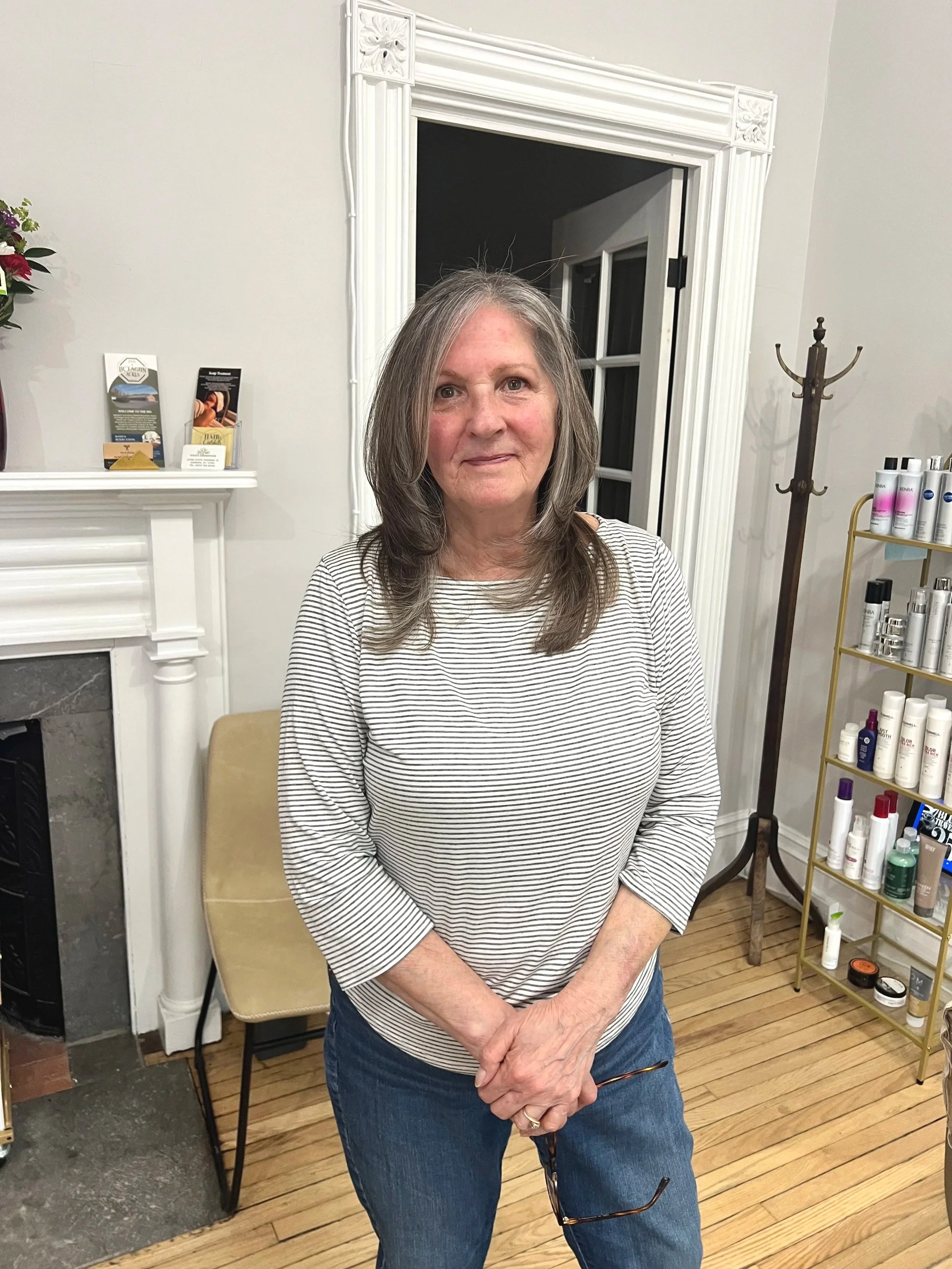 A woman with gray hair and a striped long sleeve shirt standing indoors in front of a white fireplace mantel and a wooden floor, holding glasses in her hand, with a coat rack and a shelf of skincare products nearby.