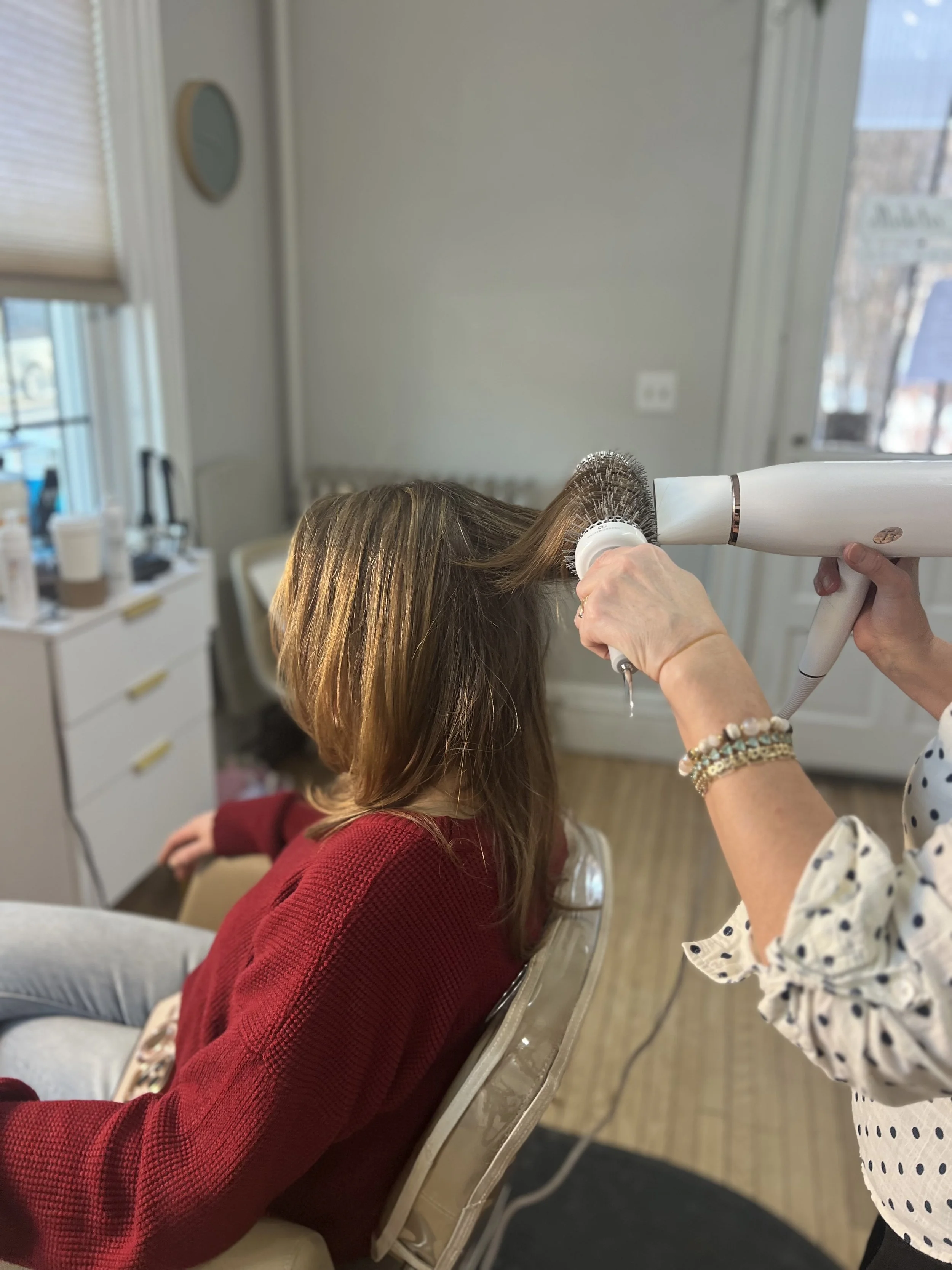 A woman drying a woman's long brown hair with a handheld hair dryer while she sits on a salon chair.