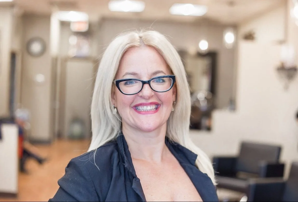 A smiling woman with blonde hair, glasses, and a dark top standing in an indoor setting resembling a salon or office.
