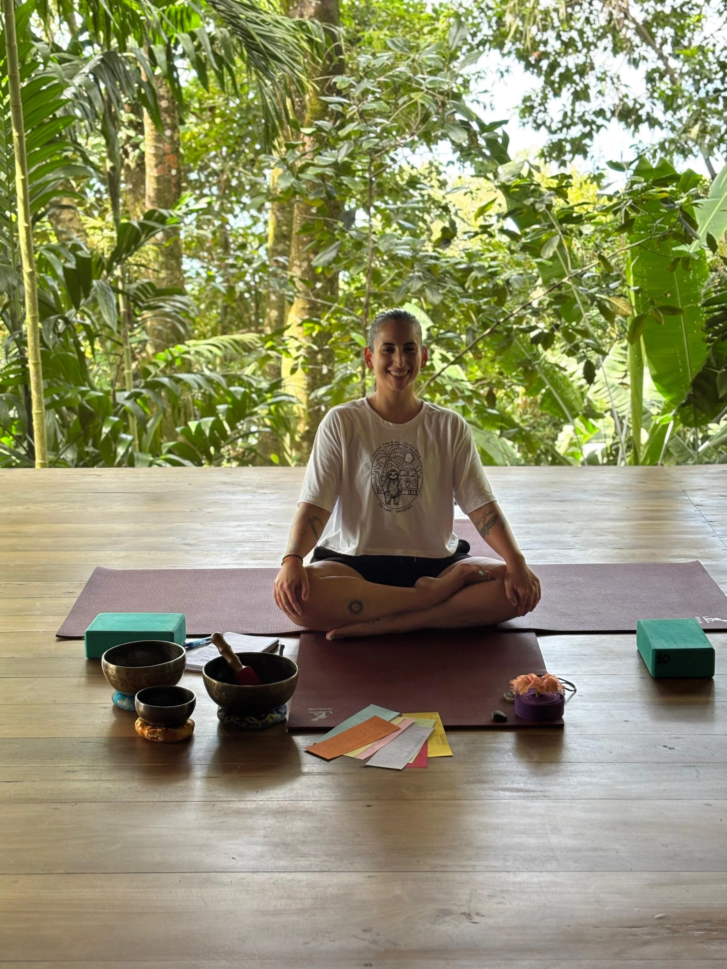 Woman sitting cross-legged on yoga mat in a lush, green outdoor setting, smiling, with singing bowls, papers, and yoga props in front of her.