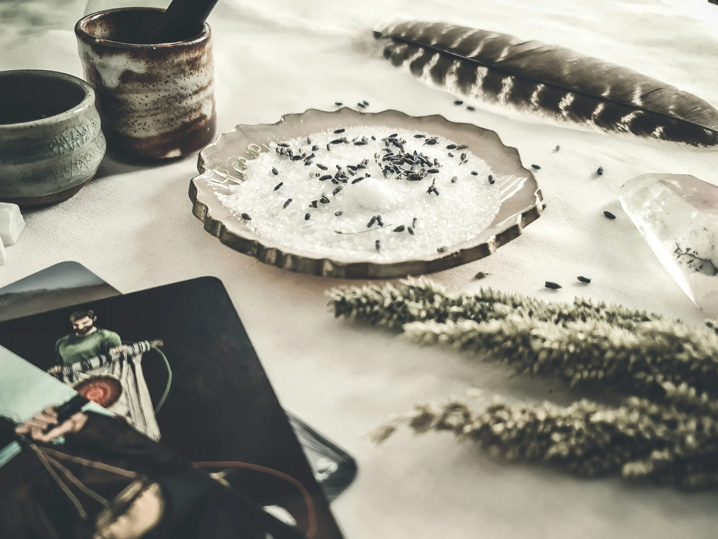 Close-up of salt and black sesame seeds in a dish, with a black feather, two small decorated jars, and a bottle of perfume on a white surface.