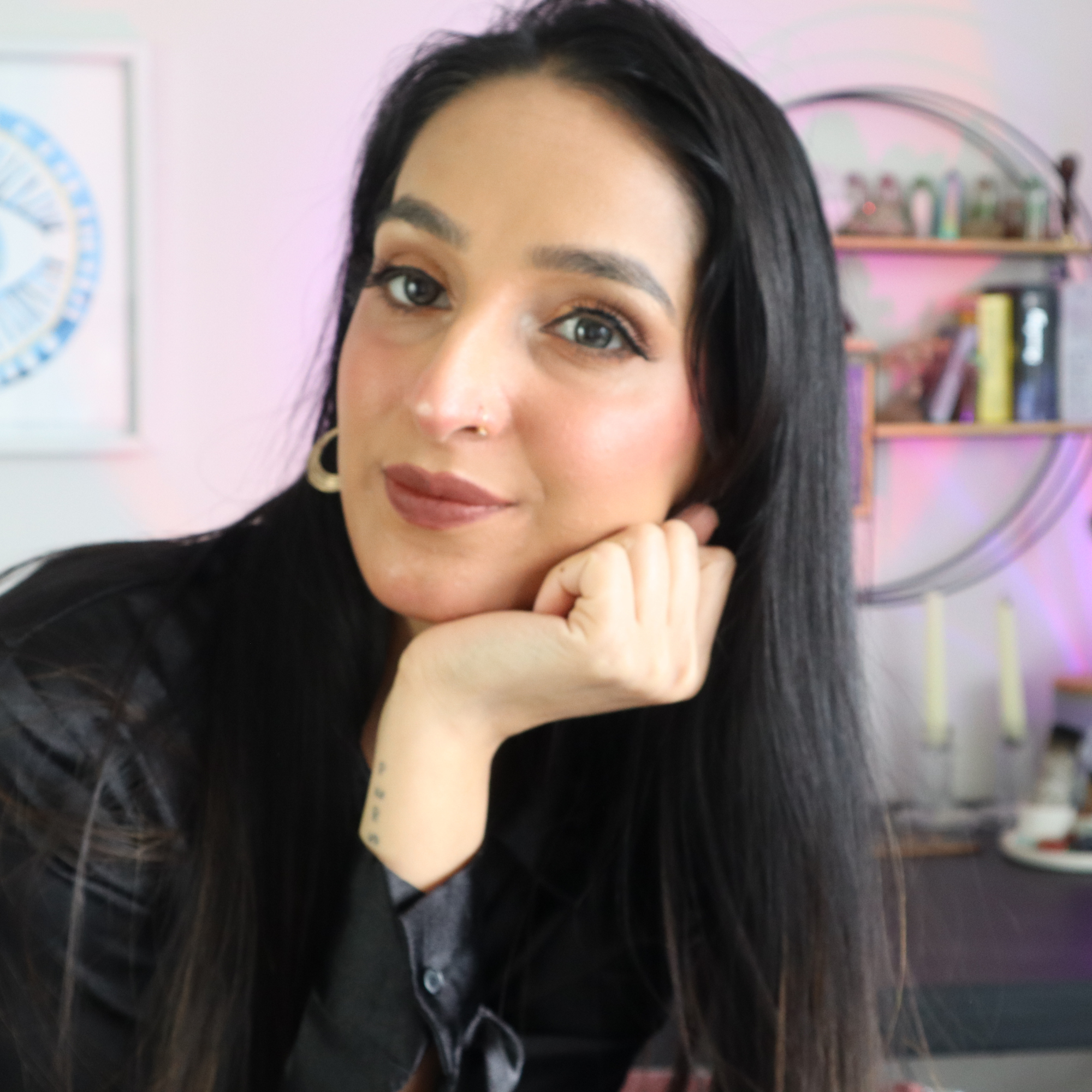 Close-up of a woman with black hair, wearing makeup, and earrings, smiling gently, with a pink wall and bookshelf in the background.