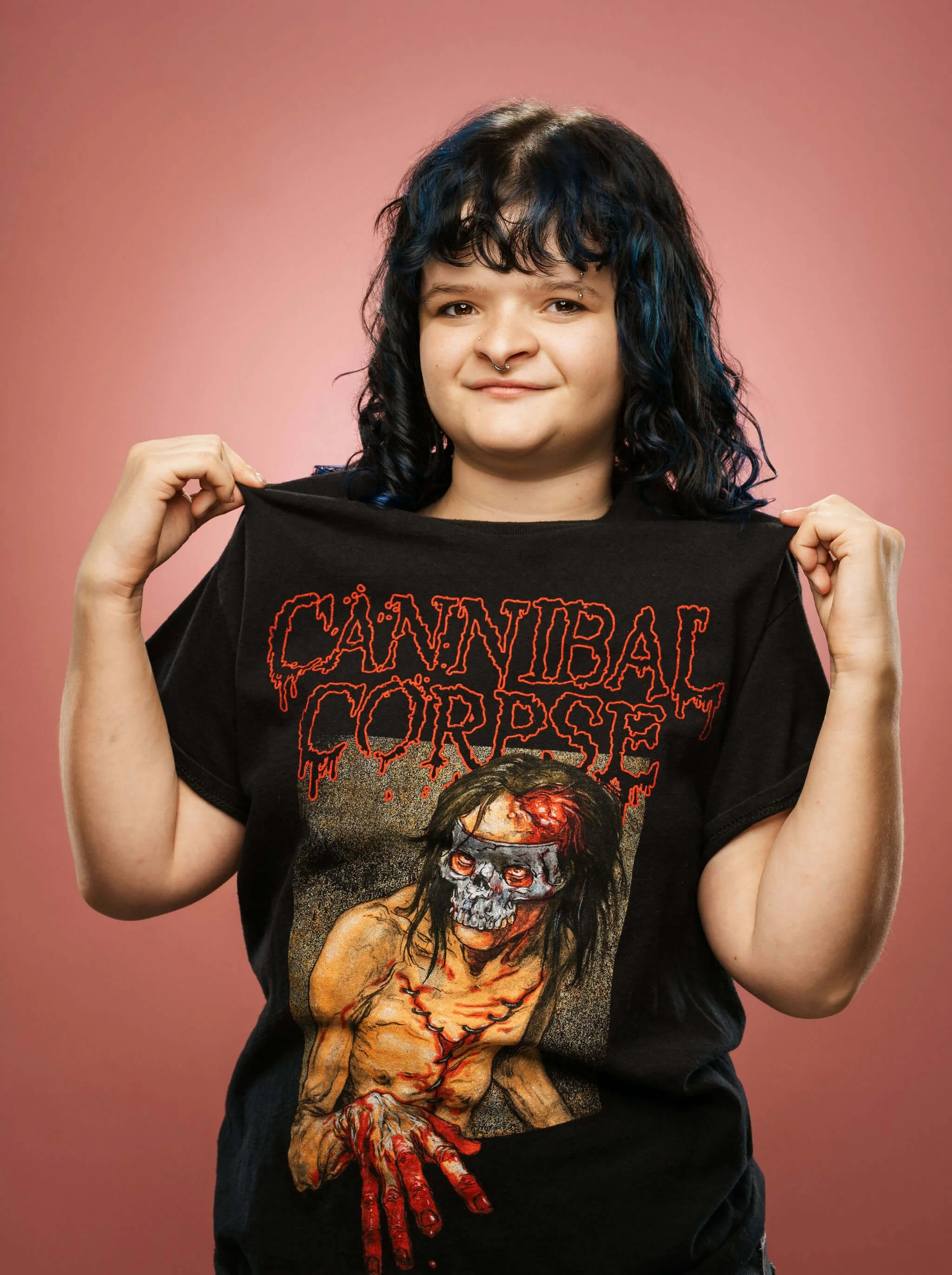 A young senior girl in high school standing in front of a pink backdrop showing off her cannibal corpse shirt