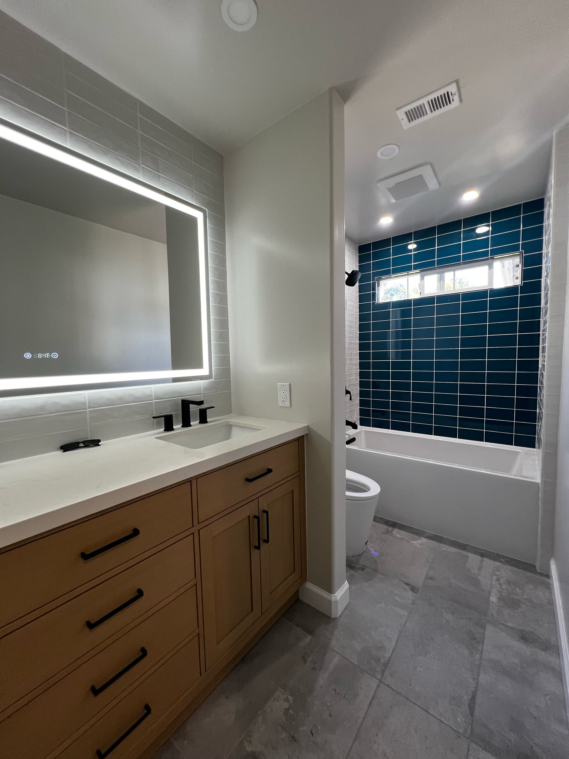 A modern bathroom featuring a large mirror with LED lighting, a wooden vanity with black handles, a white sink, a black faucet, grey tiled floors, a toilet, and a separate shower area with dark blue tiles, a small horizontal window, and a rain shower