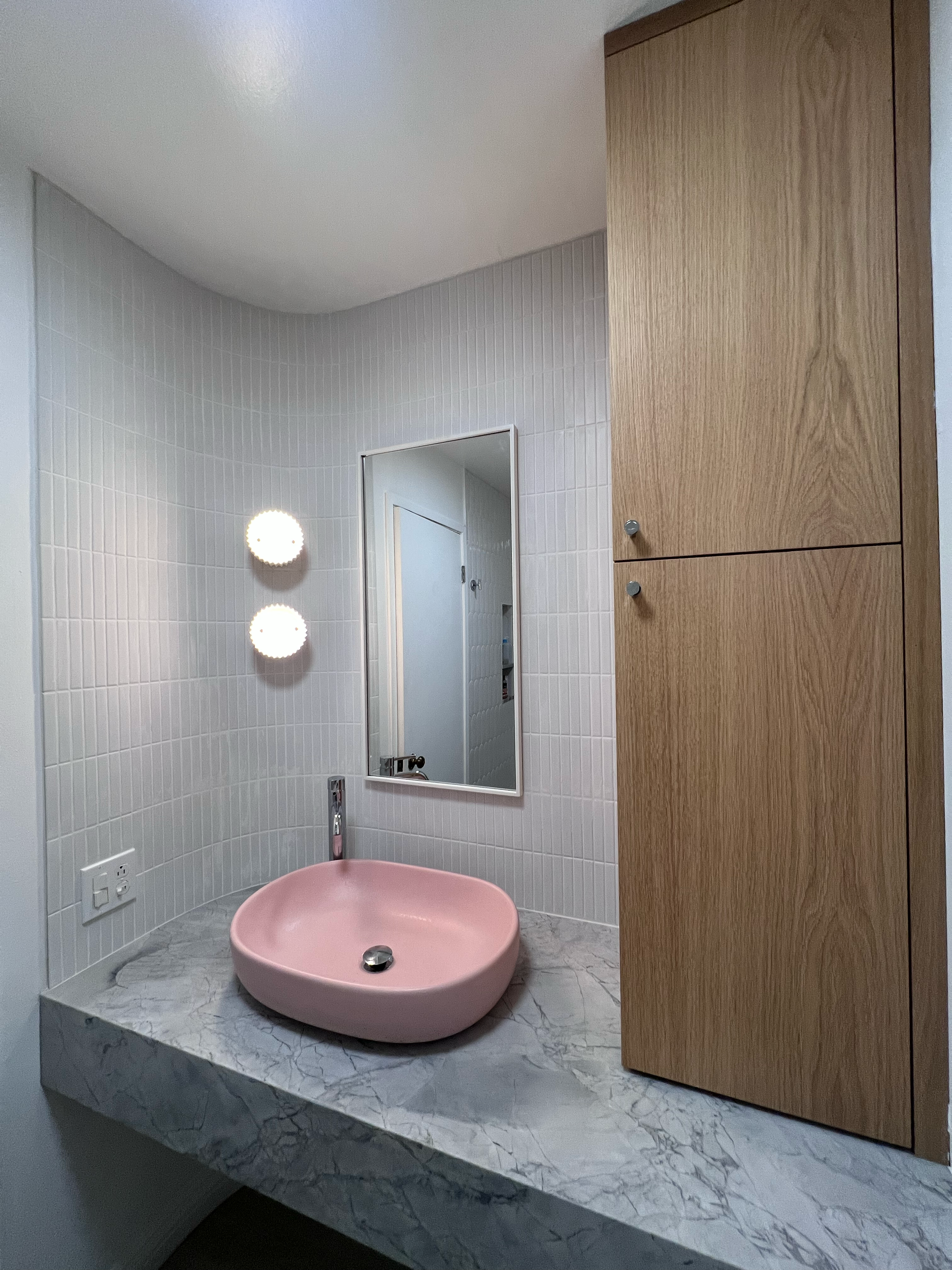 Bathroom remodel with a pink sink basin, wood cabinetry, marble countertop, round light fixtures and a white tiled wall