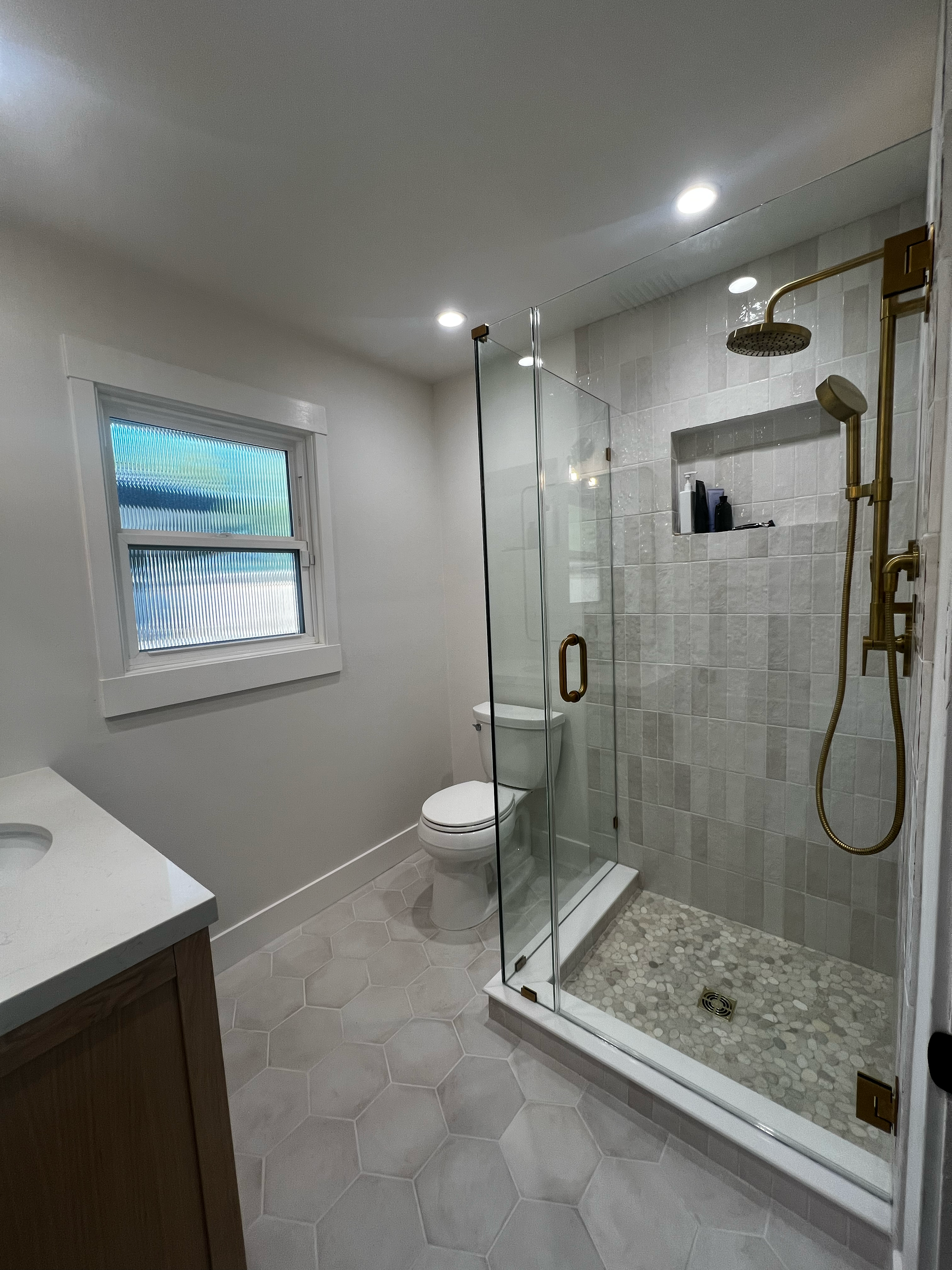 Modern bathroom with a glass shower enclosure, subway tiles, a rain shower head, handheld shower, toilet, window with textured glass, and hexagon floor tiles.