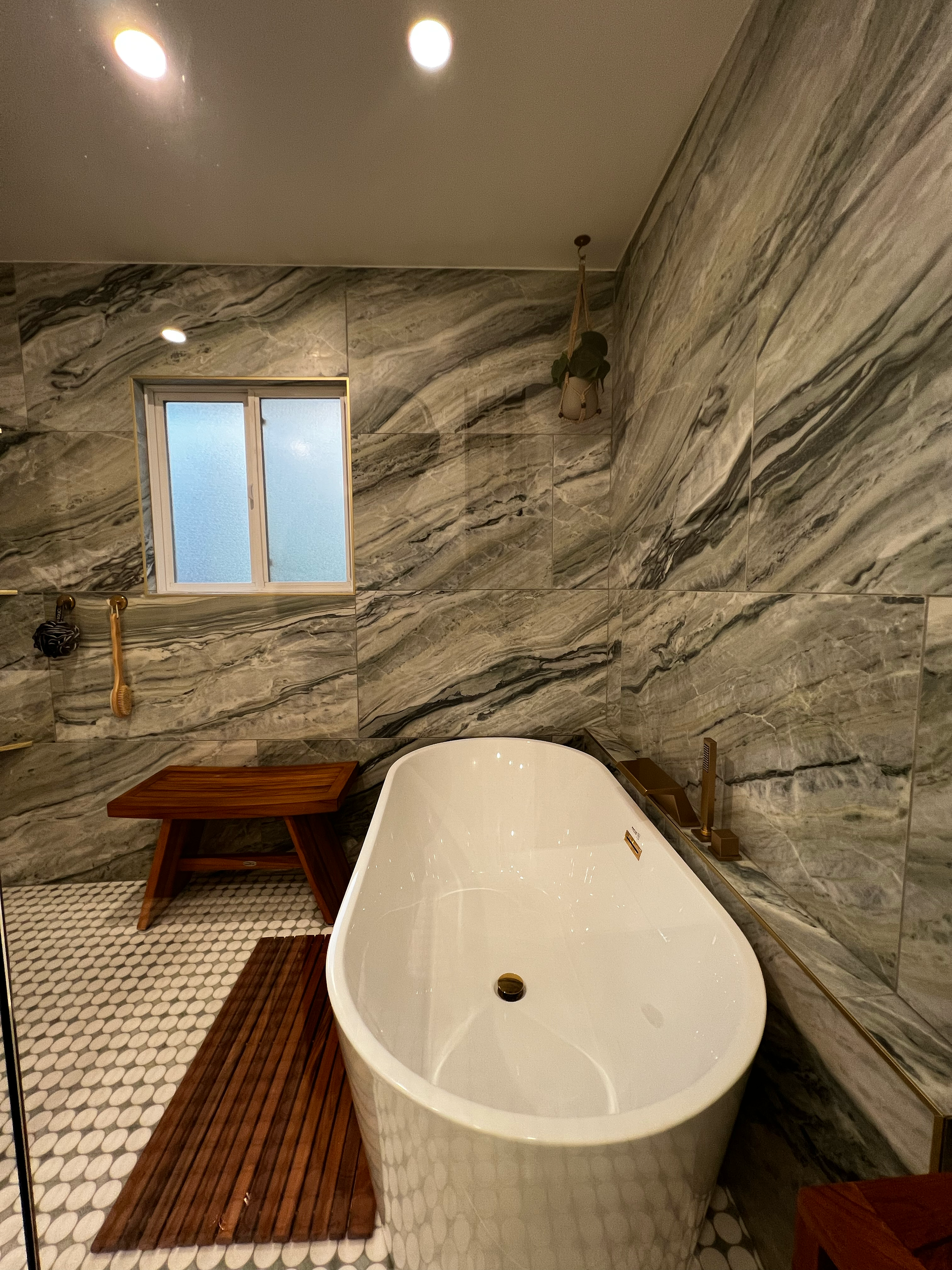 A bathroom with a white freestanding bathtub, wooden bath mat, a small window with textured glass, and marble wall tiles.