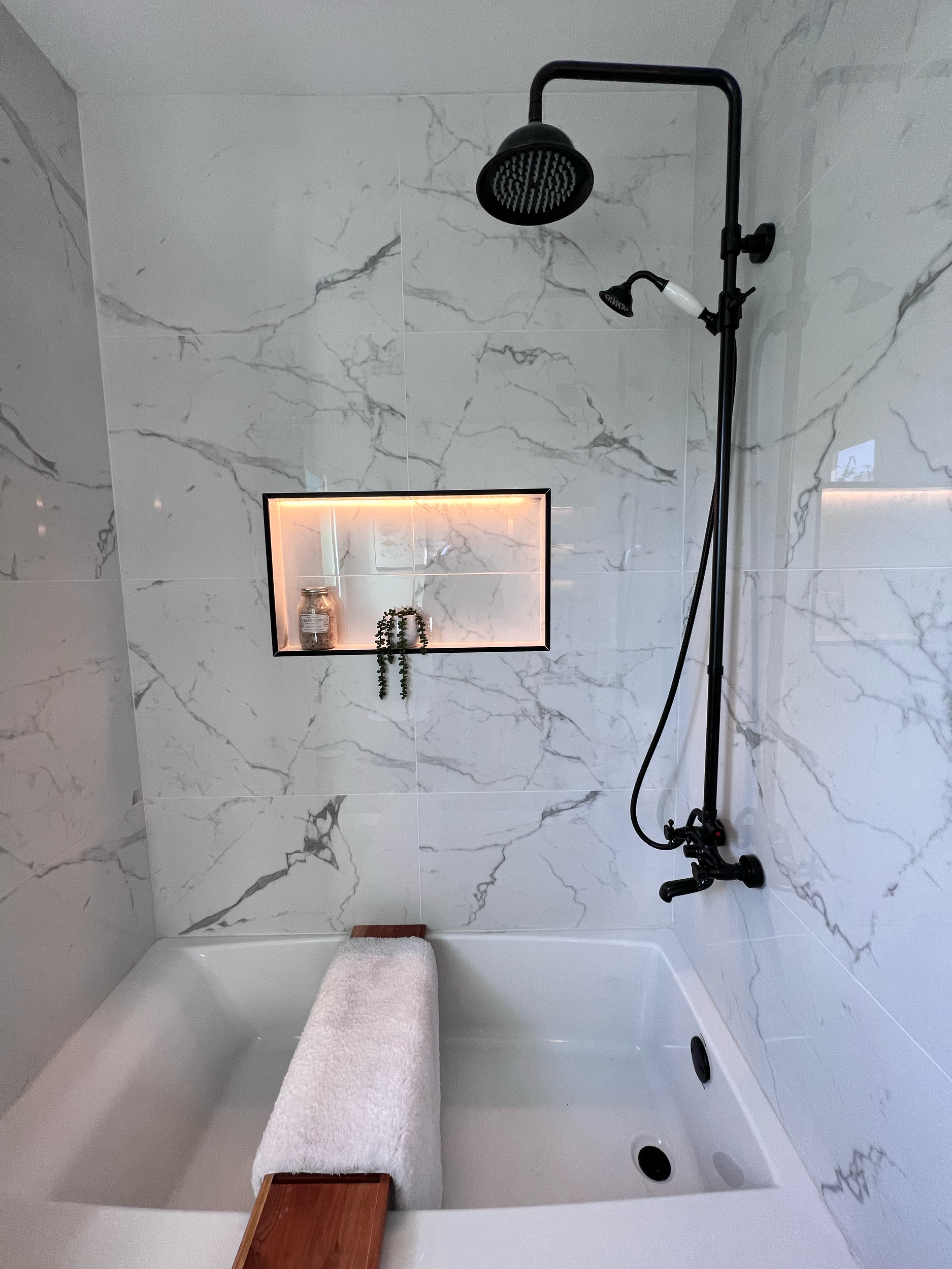A modern shower area with marble walls, a black showerhead and a handheld shower, a small recessed shelf with a jar and plant, a white towel on a wooden bench, and a partial view of a bathtub.