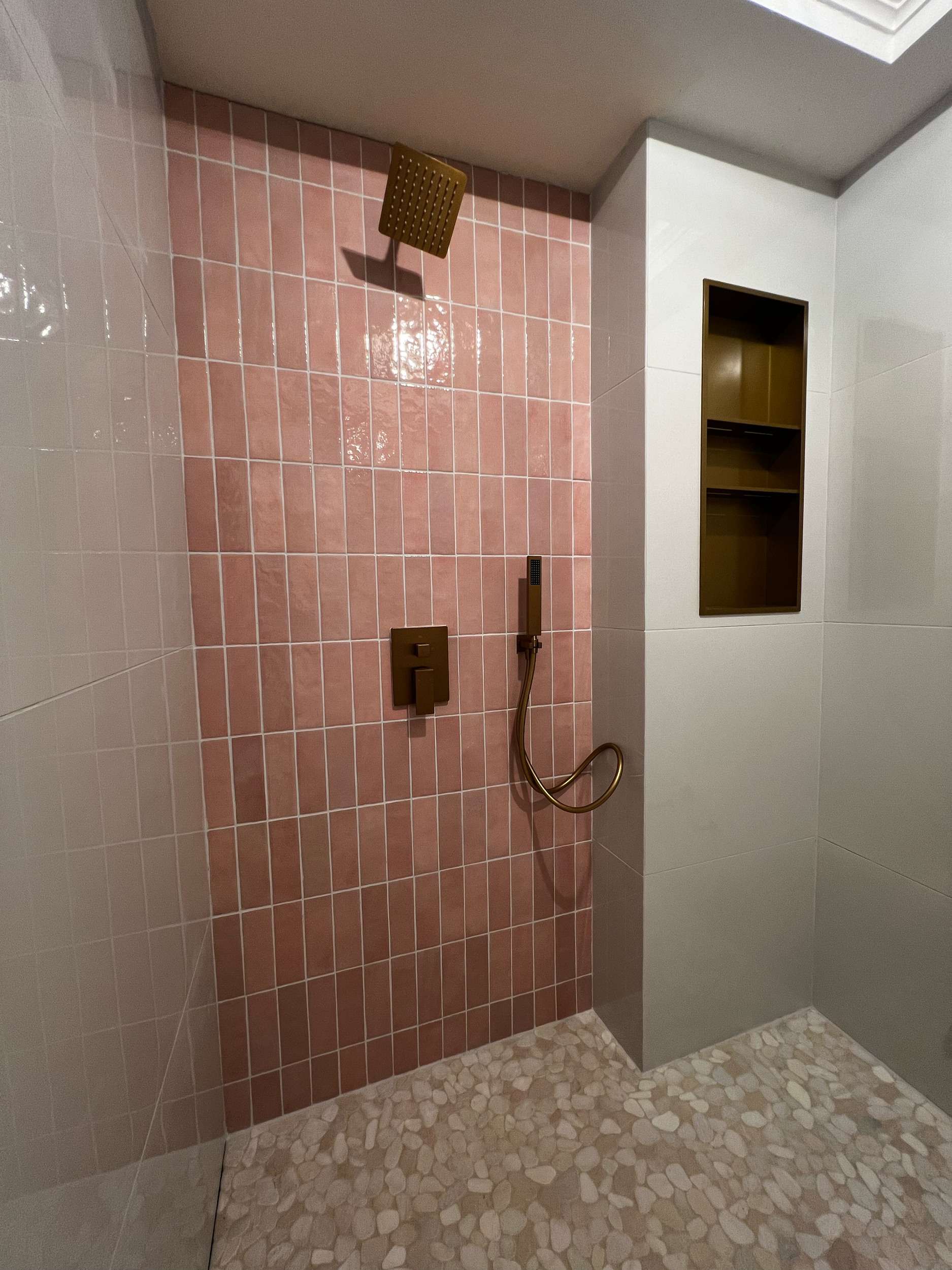 A shower area with pink tiles on the wall, a brown showerhead, and a brown handheld shower fixture hanging on the wall. The floor has a pebble pattern, and there is a small brown shelf built into the wall.