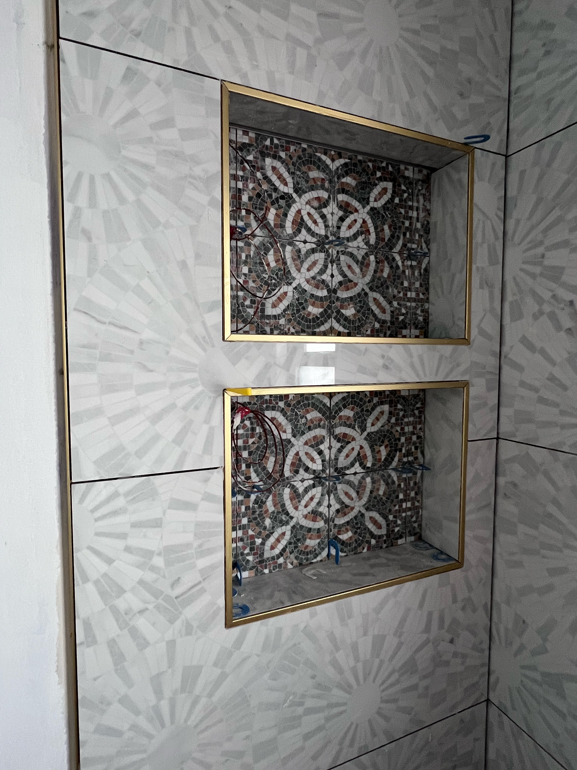 The image shows a wall with light-colored tile with a radial pattern. In the center, there are two inset areas framed with gold trim, displaying decorative mosaic tile with a floral pattern. Plumbing pipes are visible inside the inset areas, indicati