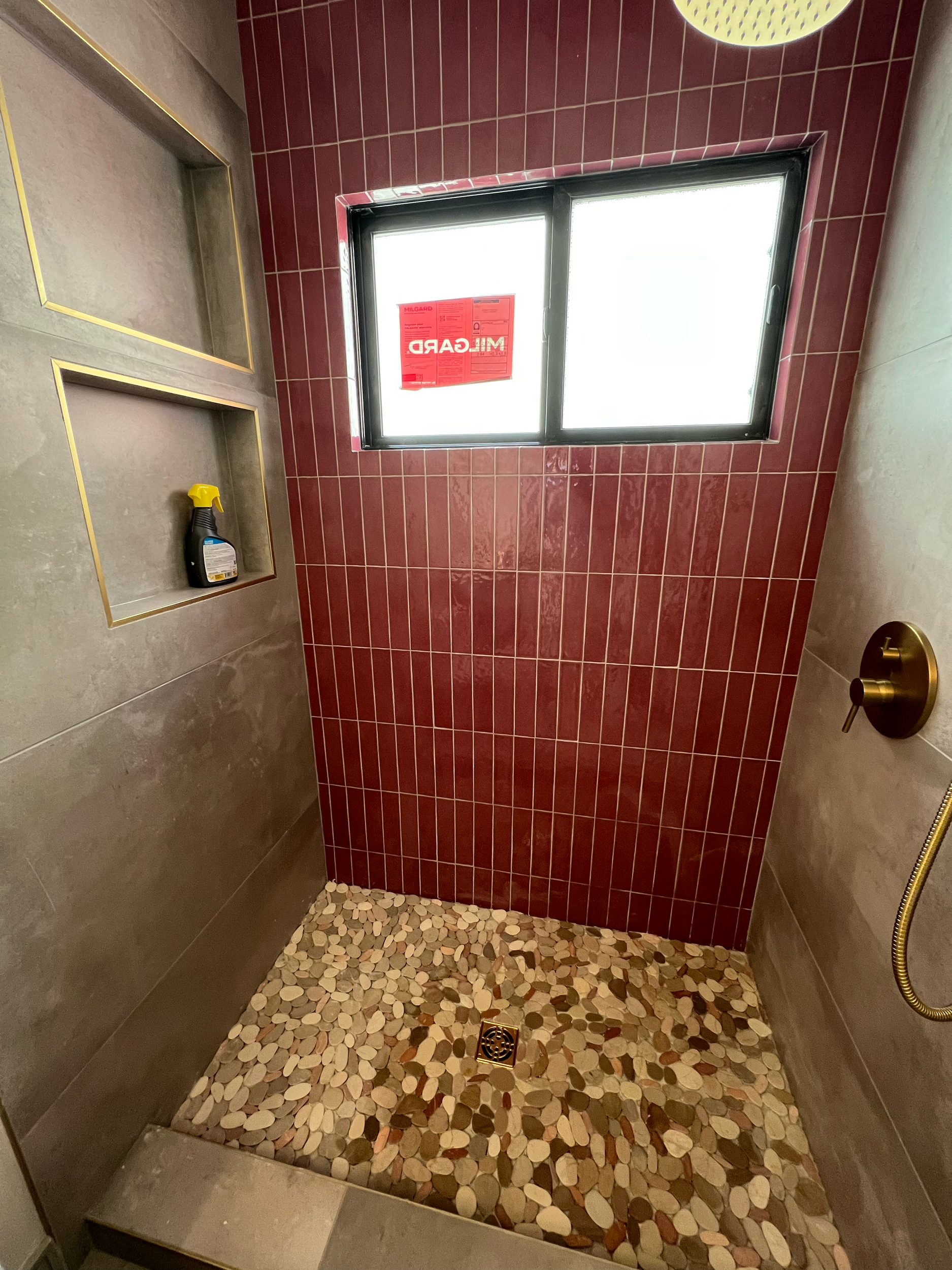 Shower area with a pebble floor, a red tiled wall, a window with a red sticker, a built-in shelf with a cleaning spray, and a gold shower handle.
