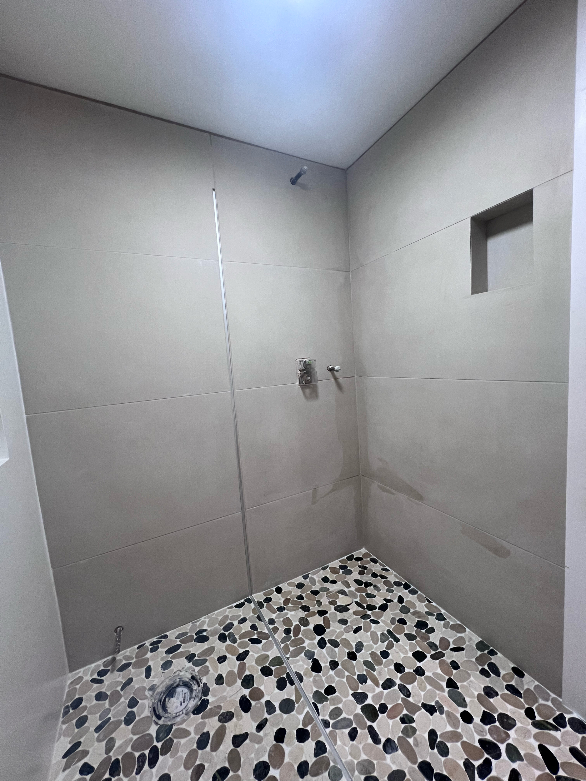 Empty walk-in shower with beige tiled walls, a small wall niche, and pebble tiled floor.