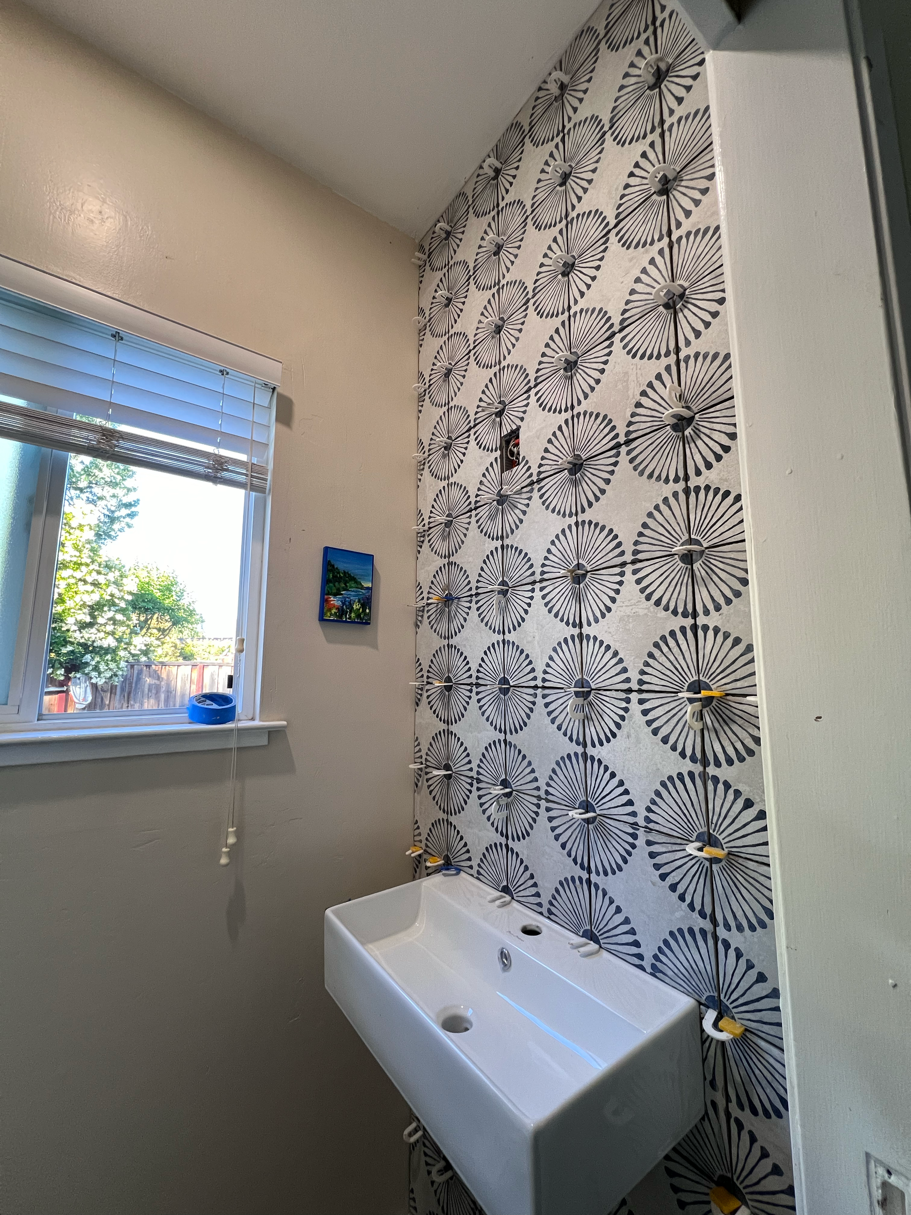 Bathroom with a window, light-colored walls, a patterned accent wall with black and white fan-shaped designs, and a white rectangular sink with no faucet. There are electrical wires and clamps on the patterned wall.